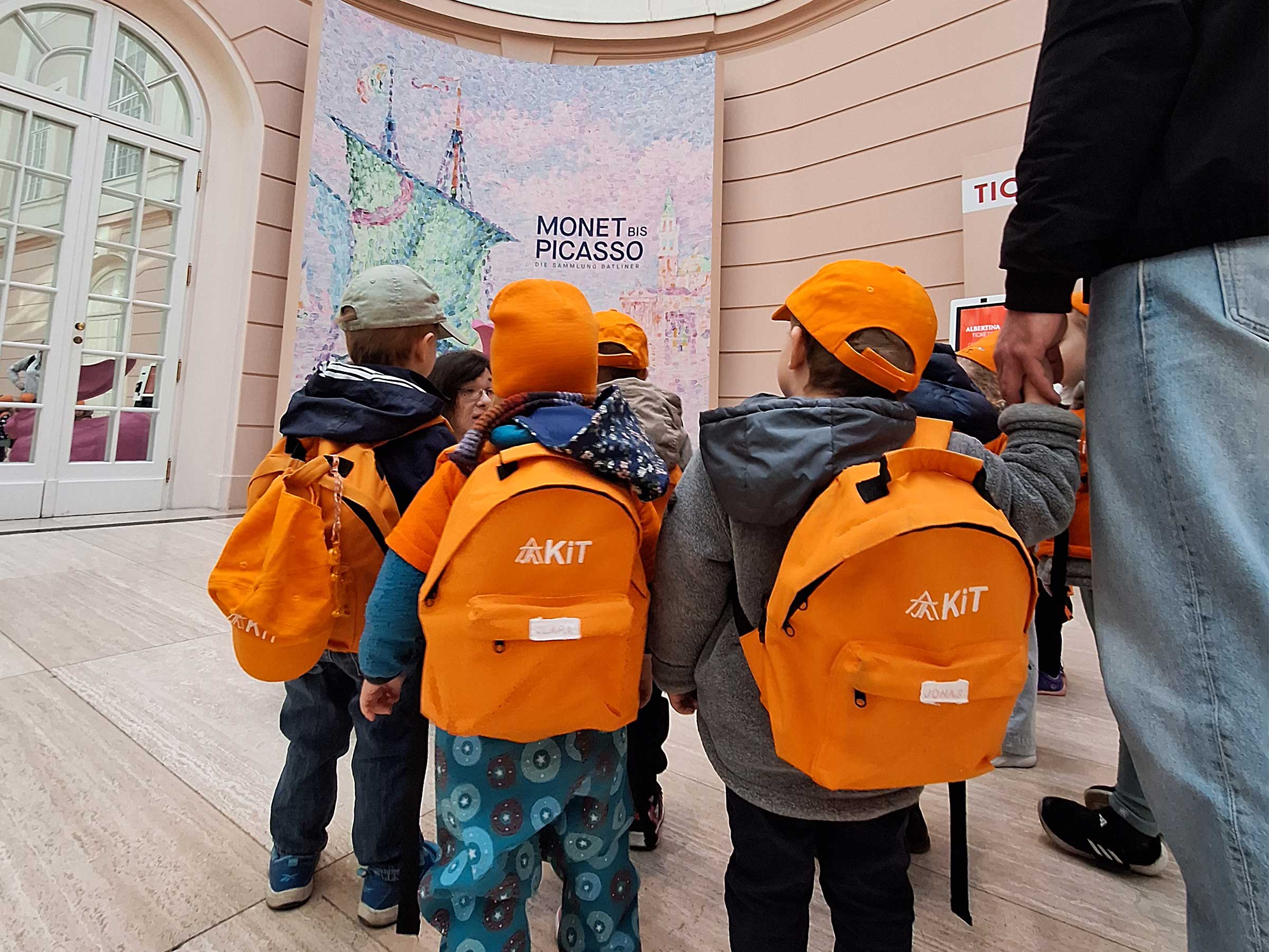 Gruppe von Kindern von hinten aufgenommen alle mit orangenen Basecaps in Richtung eines Ausstellungsschildes Monet bis Picasso gehend. Ein Kind hält die Hand einer erwachsenden Person, die nur zur Hälfte sichtbar ist.