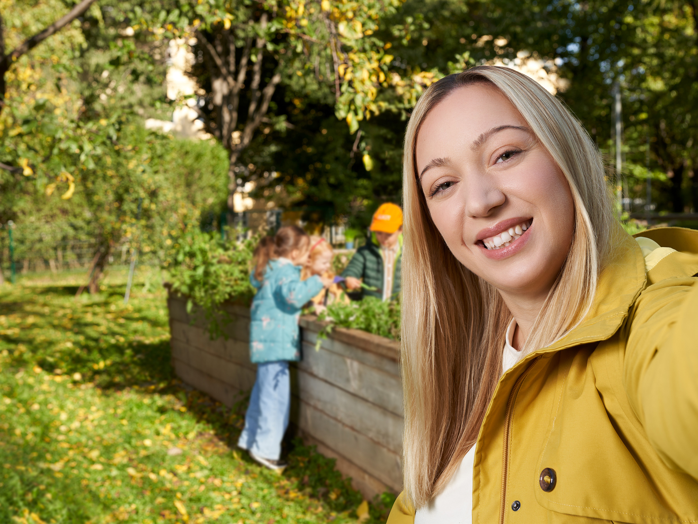 Eine blonde Frau lächelt im botanischen Garten und ist im Vordergrund sichtbar. Im Hintergrund sieht man 3 Kinde, die beim Hochbeet stehen.