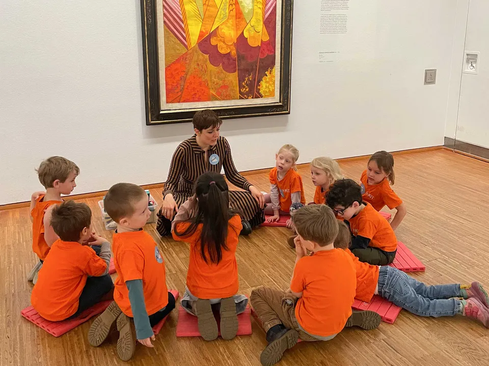Gruppe von Kindern alle in orange-farbigen T-Shirts in einem Kreis um eine erwachsene Frau sitzend vor einem großem Museumsgemälde