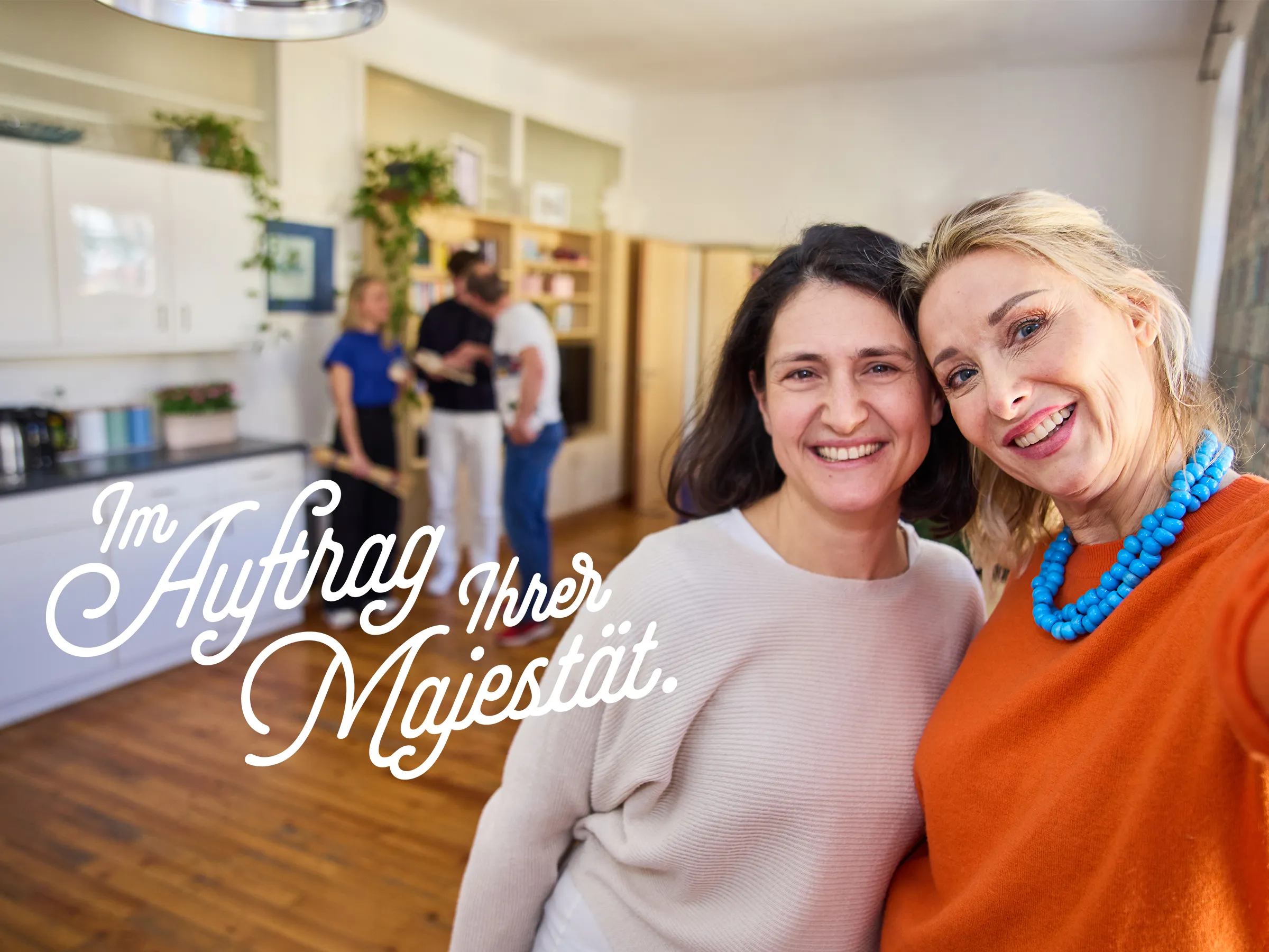 Eine Frau mit dunklen Haaren, eine Frau mit hellen Haaren und orangefarbenen Pulli mit blauer Halskette stecken die Köpfe für ein Selfie zusammen und lachen in die Kamera. Im Hintergrund eine Gruppe mit Kollegen in einem Wohn und Küchenbereich