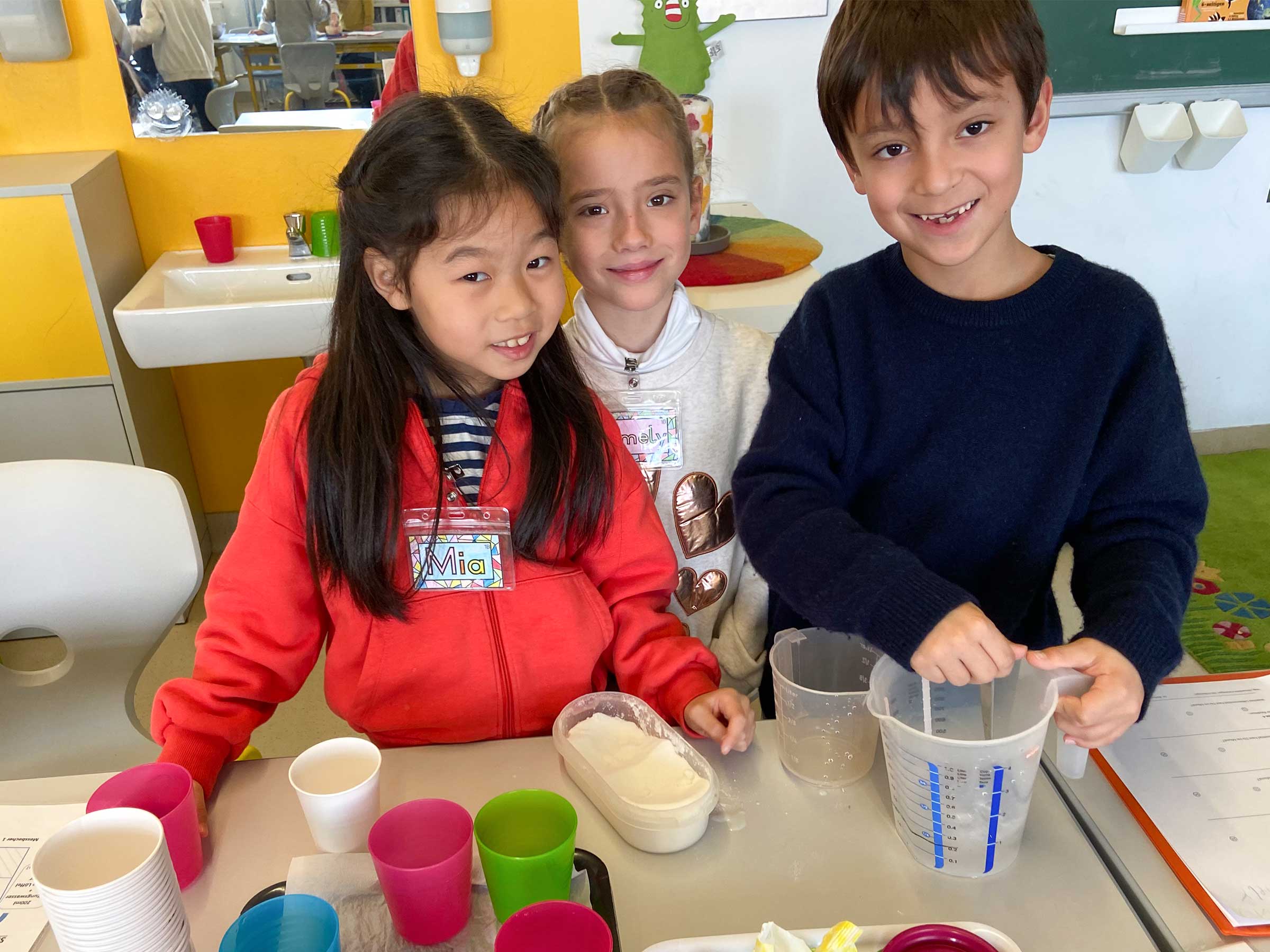 Drei Kinder an einem Tisch stehend mit bunten Bechern und einem Litermaß vor sich stehend und in die Kamera lachend.