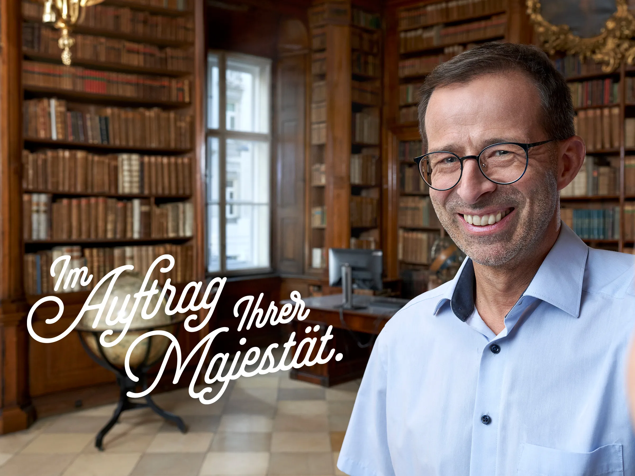 charmanter Mann mittleren Alters mit leicht schütterem Haar, Brille und Dreitagebart in blauem Hemd ein Selfie machend in einer historischen Biblitothek
