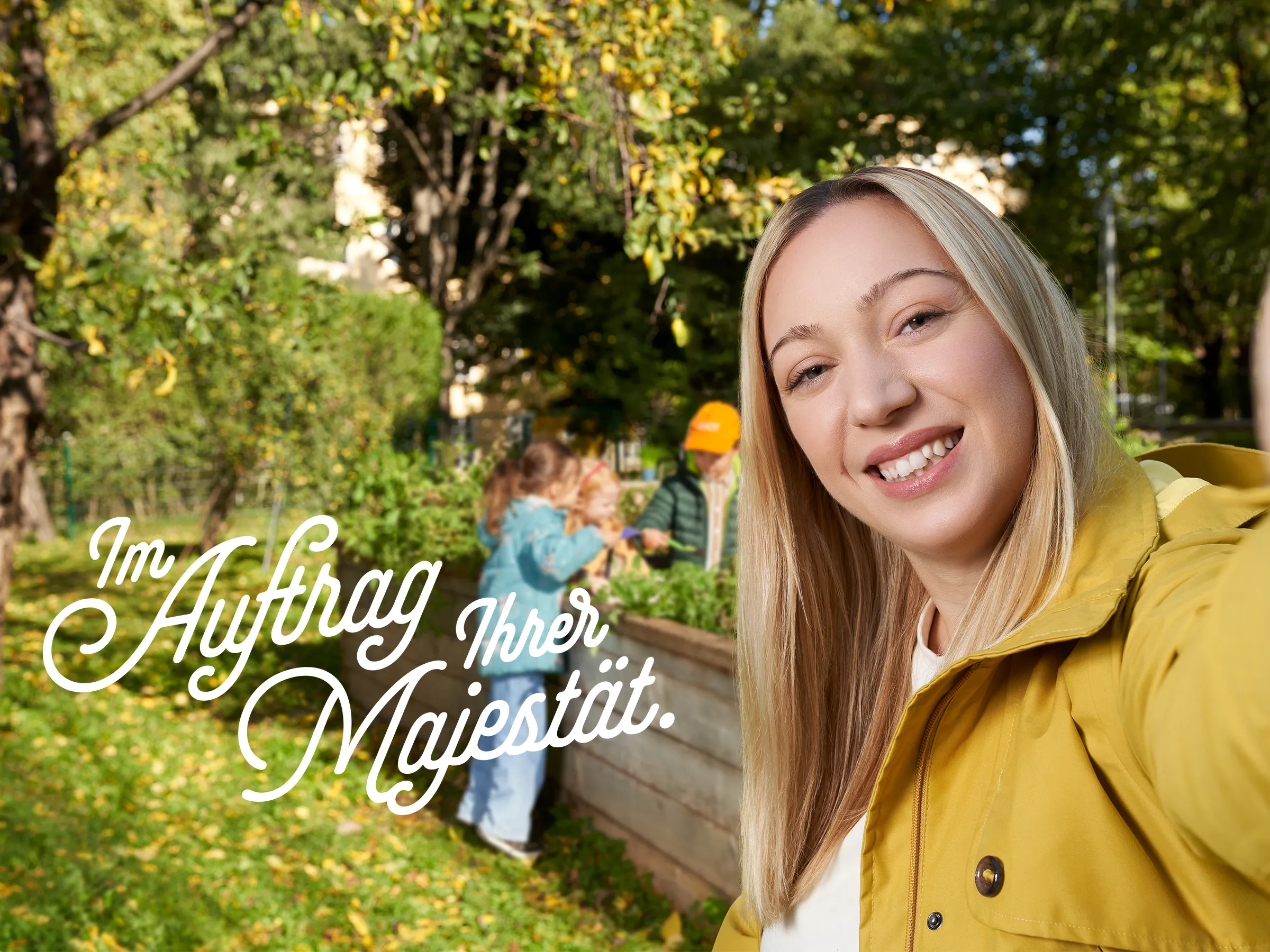 lachende junge Frau mit blonden glatten langen Haaren, gelber Outdoor Jacke ein Selfie machend mit drei Kindern im Hintergrund in einem Garten mit Hochbeet