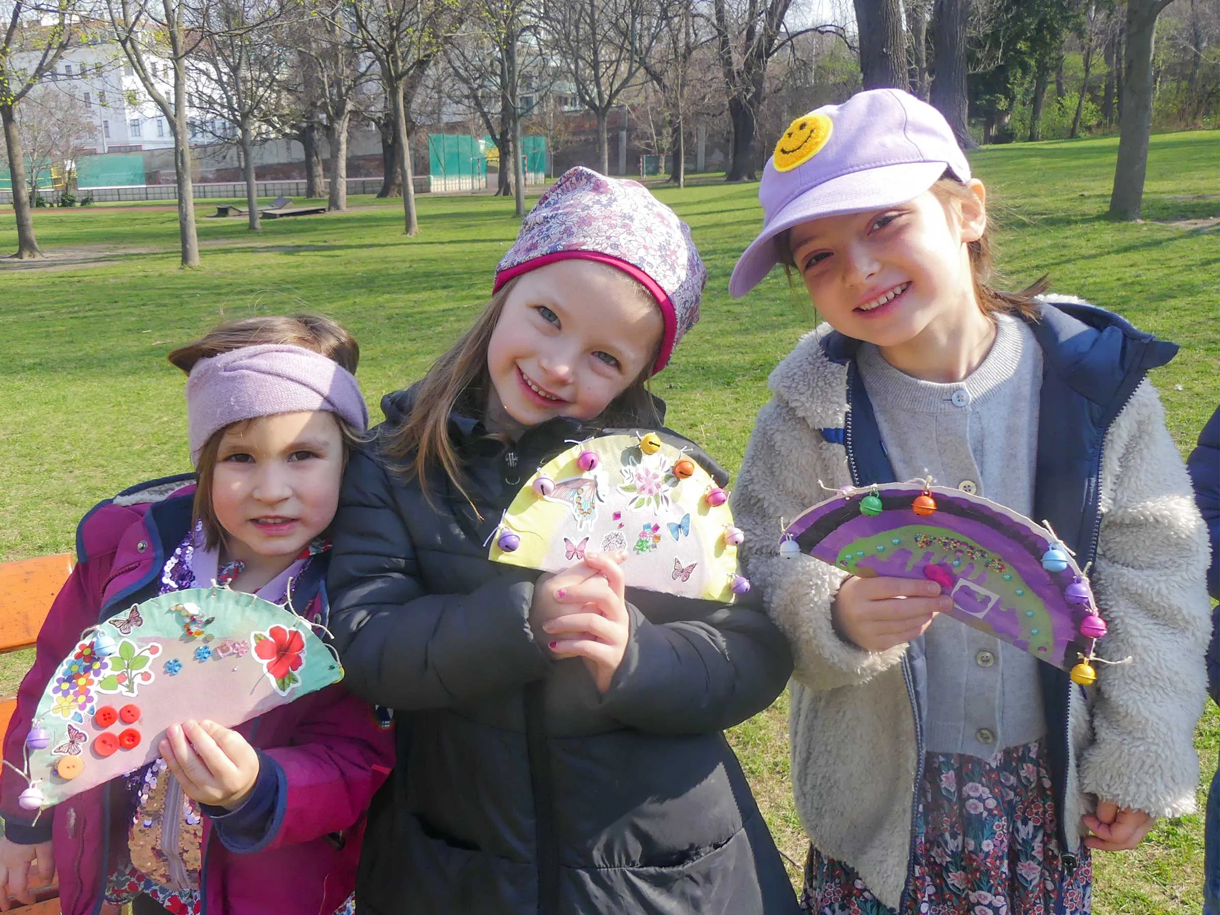 3 Mädchen in mit warmer bunter Kleidung und Mützen halten selbst gebastelte und verzierte Fächer in der Hand. Sie stehen auf einer Wiese mit Bäumen im Hintergrund.