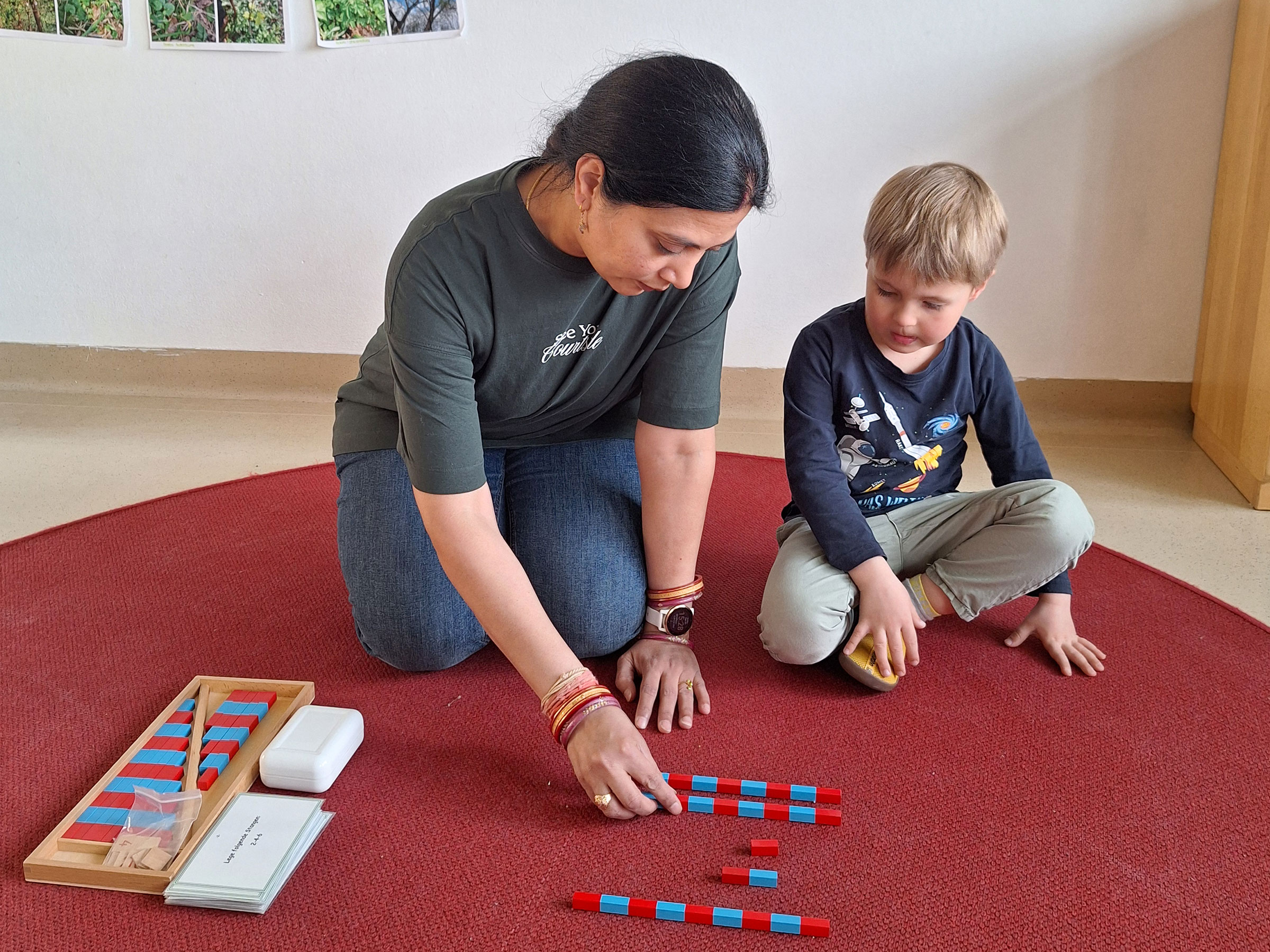 Erwachsene Frau mit kleinem Buben auf einem roten Teppichboden sitzend und mit rot blauen Bausteinen arbeitend.