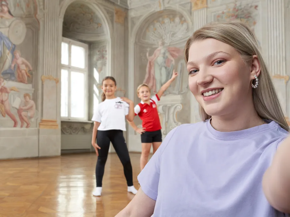 Lachende junge Frau im lila T-Shirt mit zwei Kindern in Tanspose im Hintergrund im barocken Umfeld