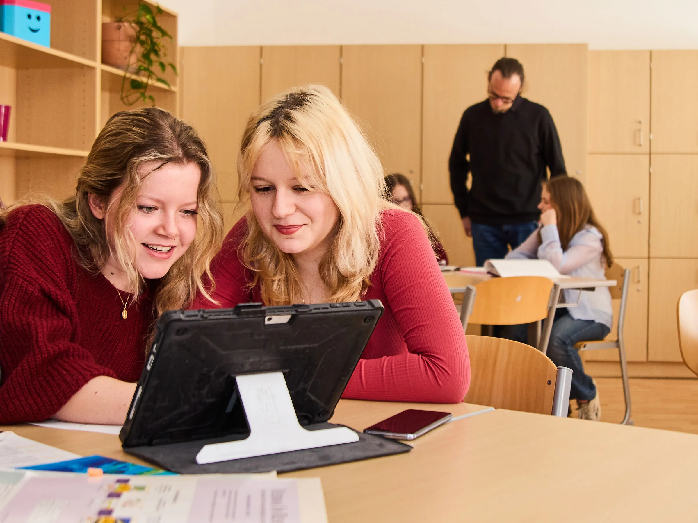 Zwei Teenager mit blonden langen Haaren an einem Tisch an einem Tablet sitzend mit eine erwachsenen Mann im Hintergrund auf zwei weitere Mädchen blickend.