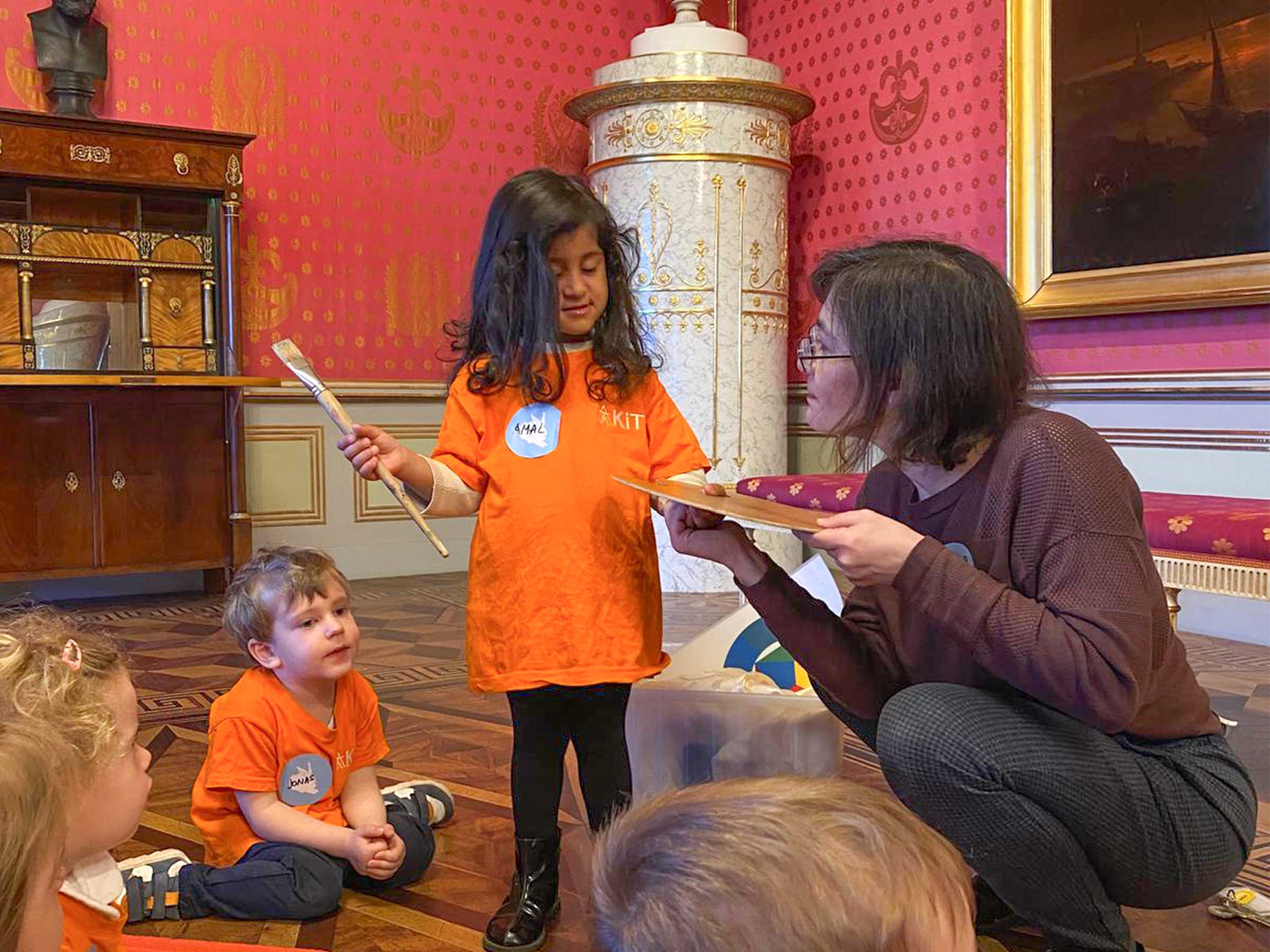 Mädchen mit orange farbigen T-Shirt und einem großem Pinsel in der Hand neben einer hockenden Frau stehend mit weiteren Kindern am Boden sitzend vor einem barocken Hintergrund