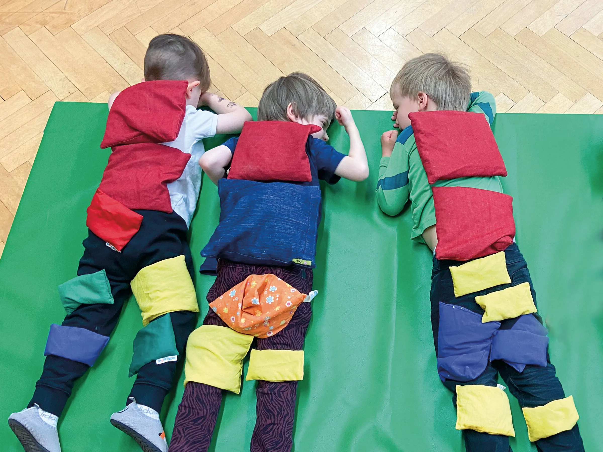 Drei Buben am Bauch auf einer grünen Turnmatte liegend bedeckt mit bunten Sandsäckchen.