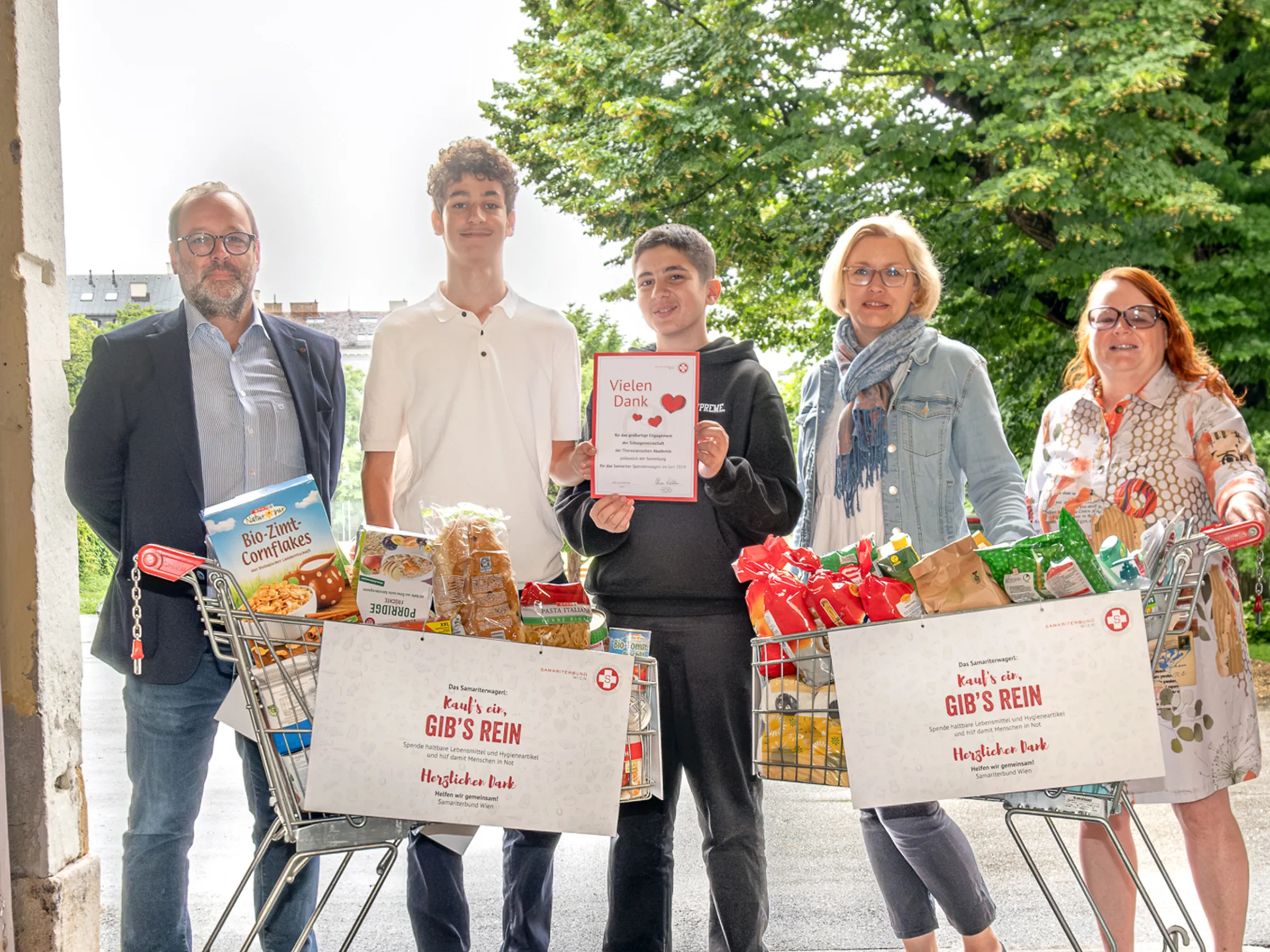 Gruppe von drei Erwachsenen und zwei Burschen mit vollen Einkaufswagerl bei frühlingshaftem sonnigen Wetter eine Spendenurkunde in die Höhe haltend