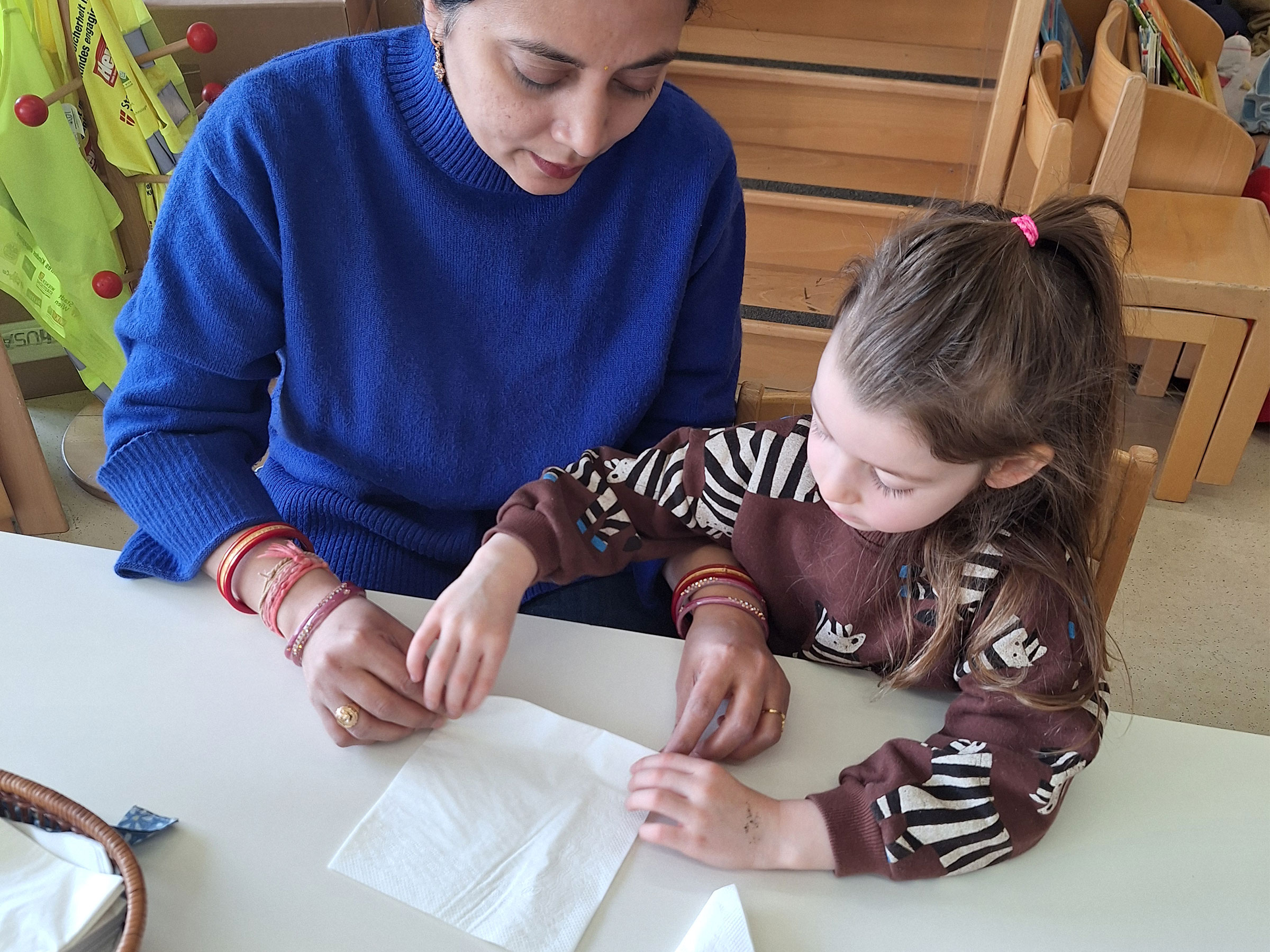 Eine Frau in blauem Pullover mit einem kleinem Mädchen an einem Tisch sitzend und eine Serviette faltend.