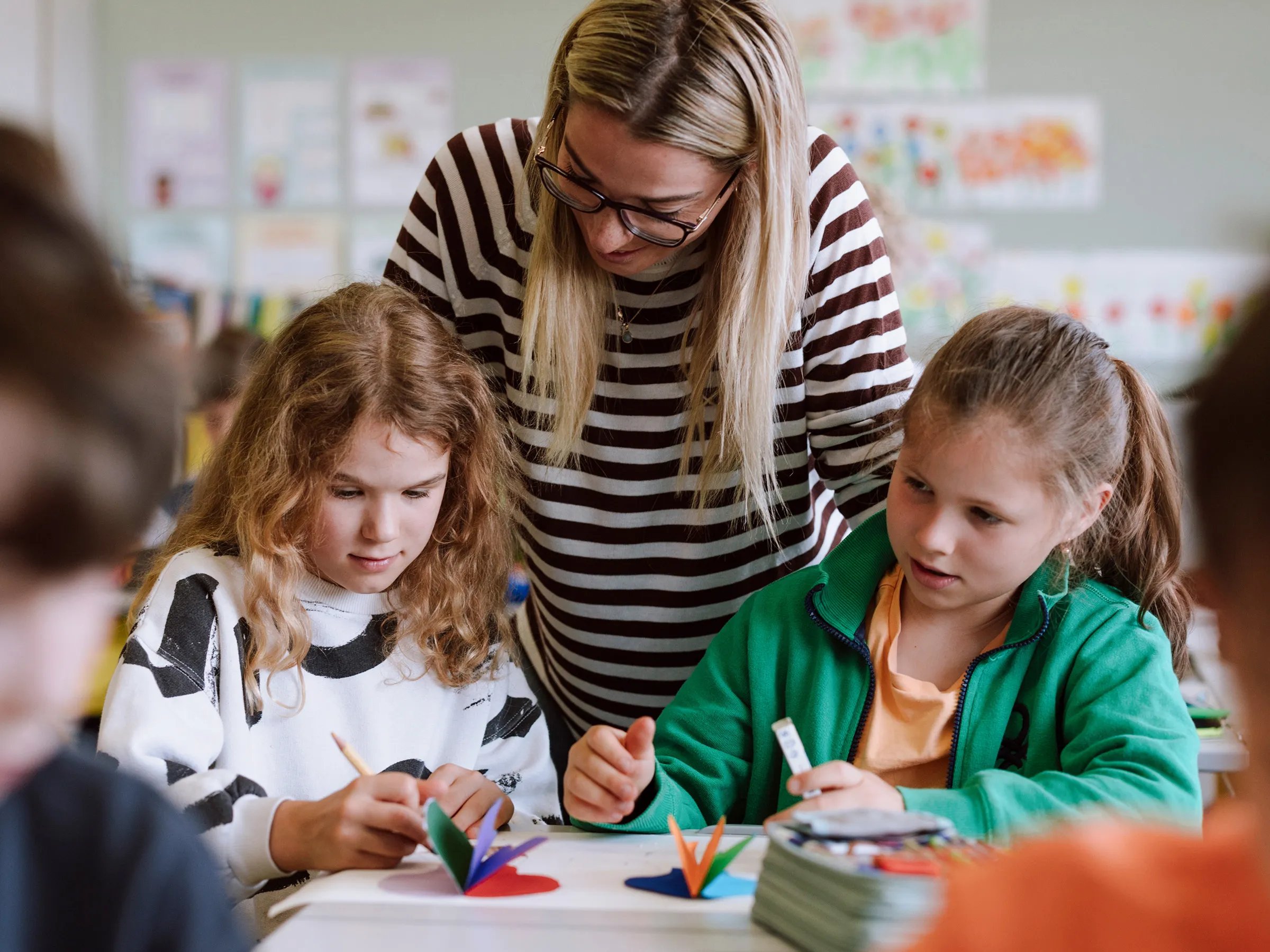 Zwei Mädchen mit langen Haaren und farbenfroh gekleidet in einer Schulbank sitzend und arbeitend mit einer jungen Lehrerin mit Brille und langen Haaren und geringeltem Pulli zwischen ihnen stehend und auf die Arbeit der Kinder blickend