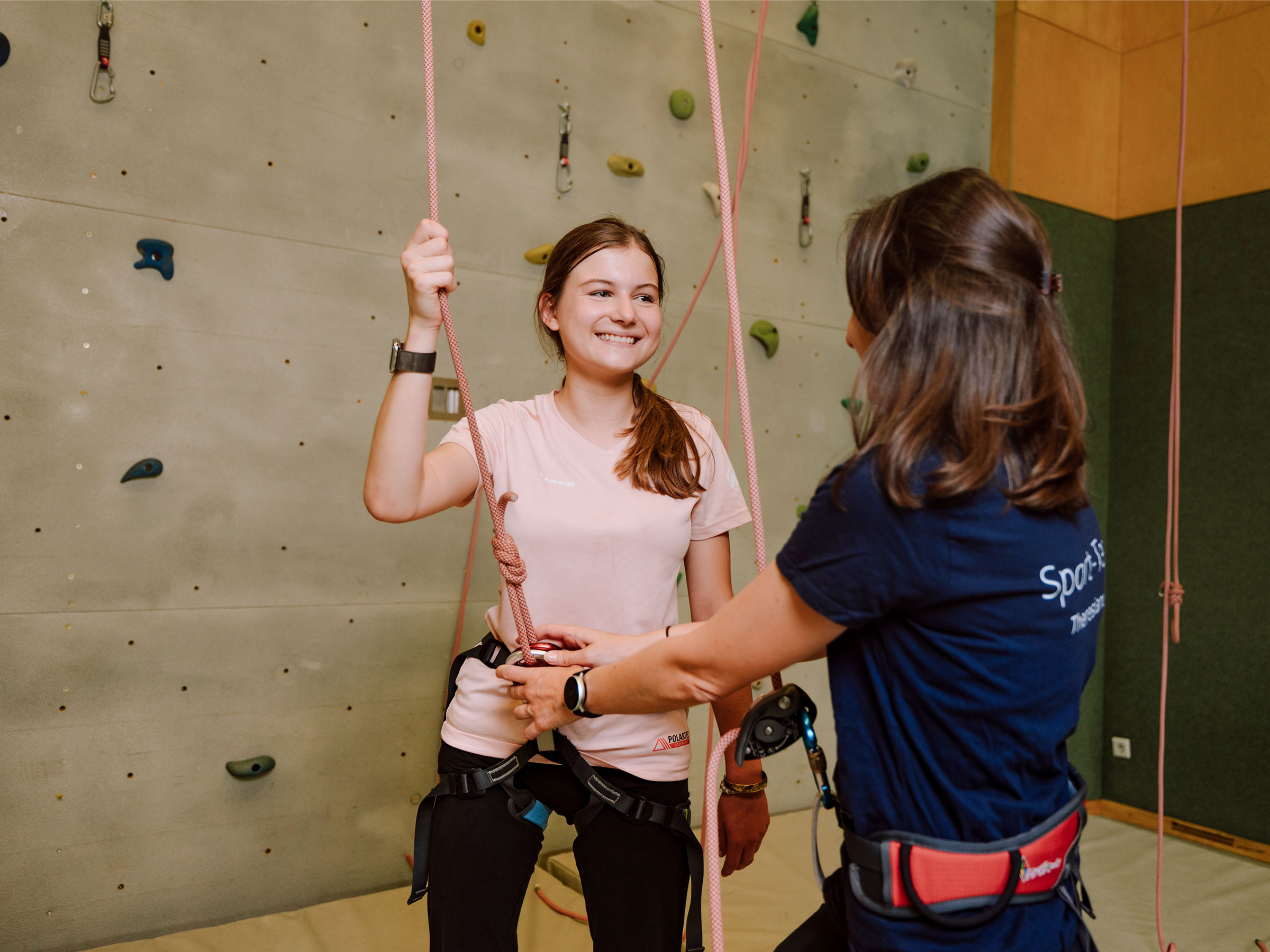 Mädchen mit langen brünetten Haaren an einer Kletterwand stehend und eine Frau anlachend, den Klettergurt prüfend.
