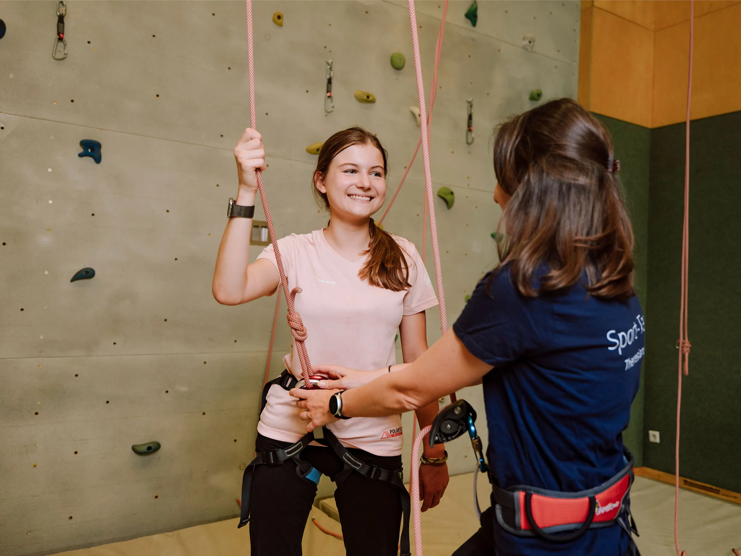 Mädchen mit langen brünetten Haaren an einer Kletterwand stehend und eine Frau anlachend, den Klettergurt prüfend.