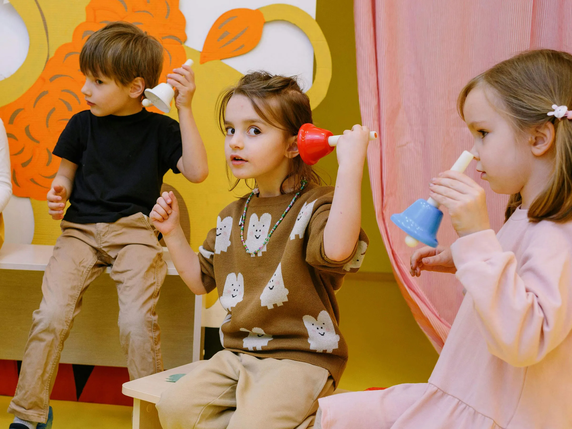 Zwei Mädchen und eine Bube mit Glocken in den Händen und neugierig dem Glockenklang lauschend.