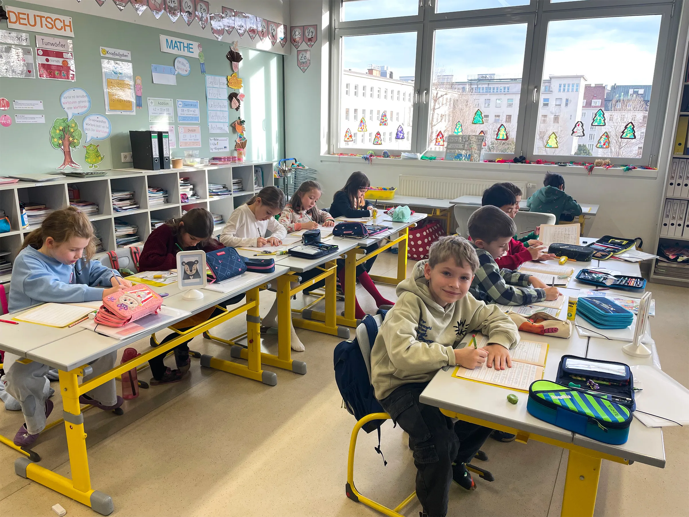 Lichtdurchflutetes Klassenzimmer mit zehn Kindern an Tischen sitzend, ein Bursche von ihnen lacht in die Kamera während er mit einem Bleistift in ein Heft schreibt