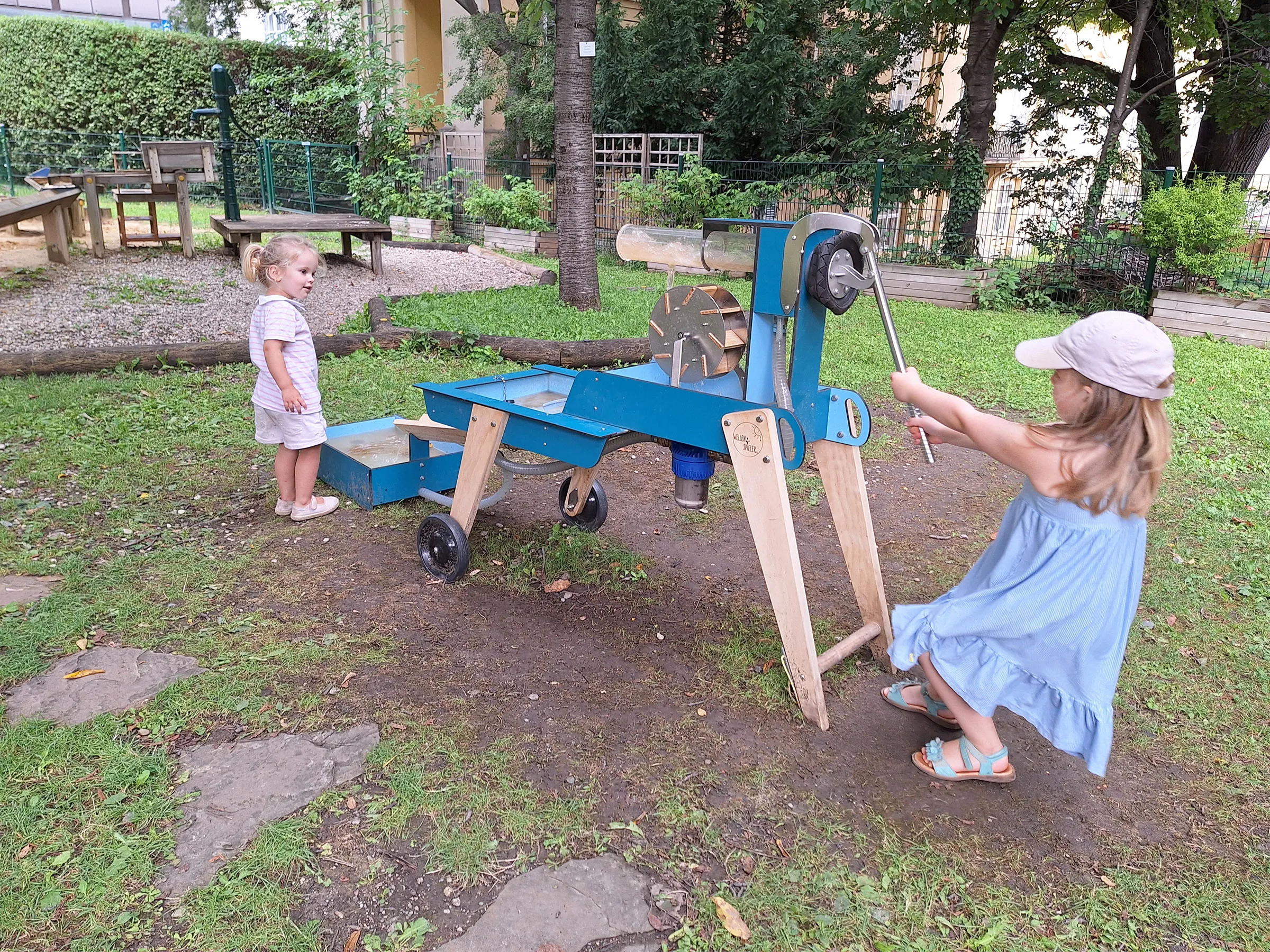 Zwei kleine Mädchen in luftiger sommerlicher Kleidung auf einem Spielplatz mit einer Wasserpumpe und einem Wasserrad beschäftigt