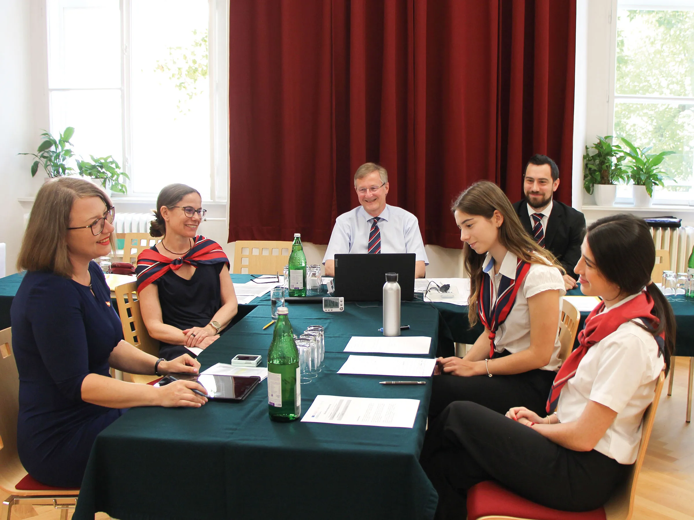 Gruppe von Männern und Frauen formell aber freundlich lachend um einen Tisch sitzend.