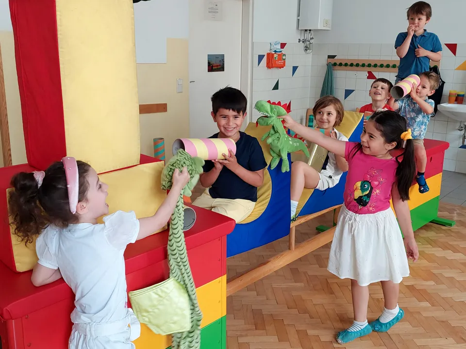 Gruppe von Vorschulkindern auf übergroßen bunten Bauklötzen sitzend und mit Stofftieren und Kartonröhren herumalbernd.