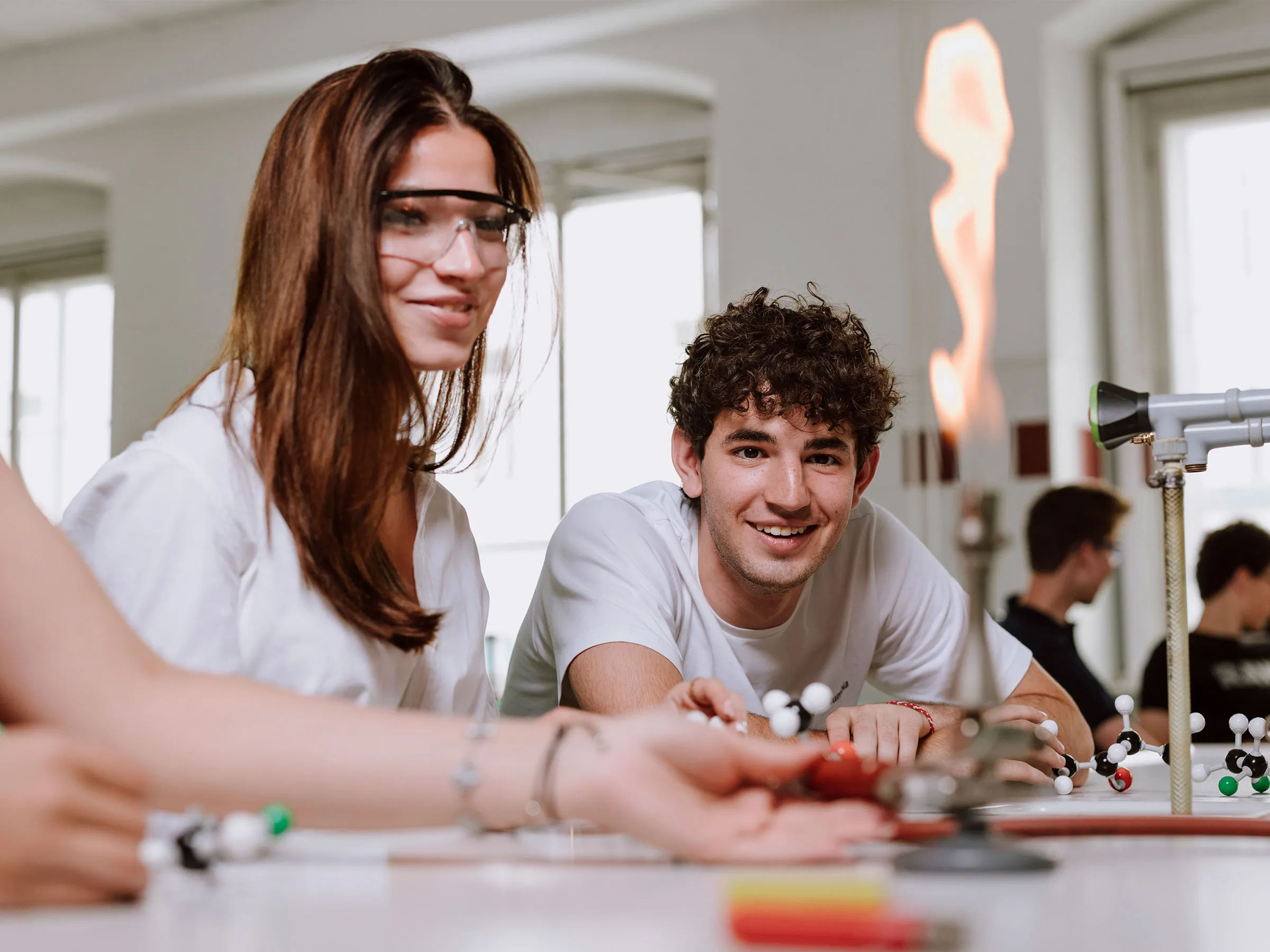Eine attraktive junge Frau mit brünetten langen Haaren und einer Sicherheitsbrille und ein interessiert schauender junger Mann mit dunklen Locken in Richtung eines Bunsenbrenners blickend
