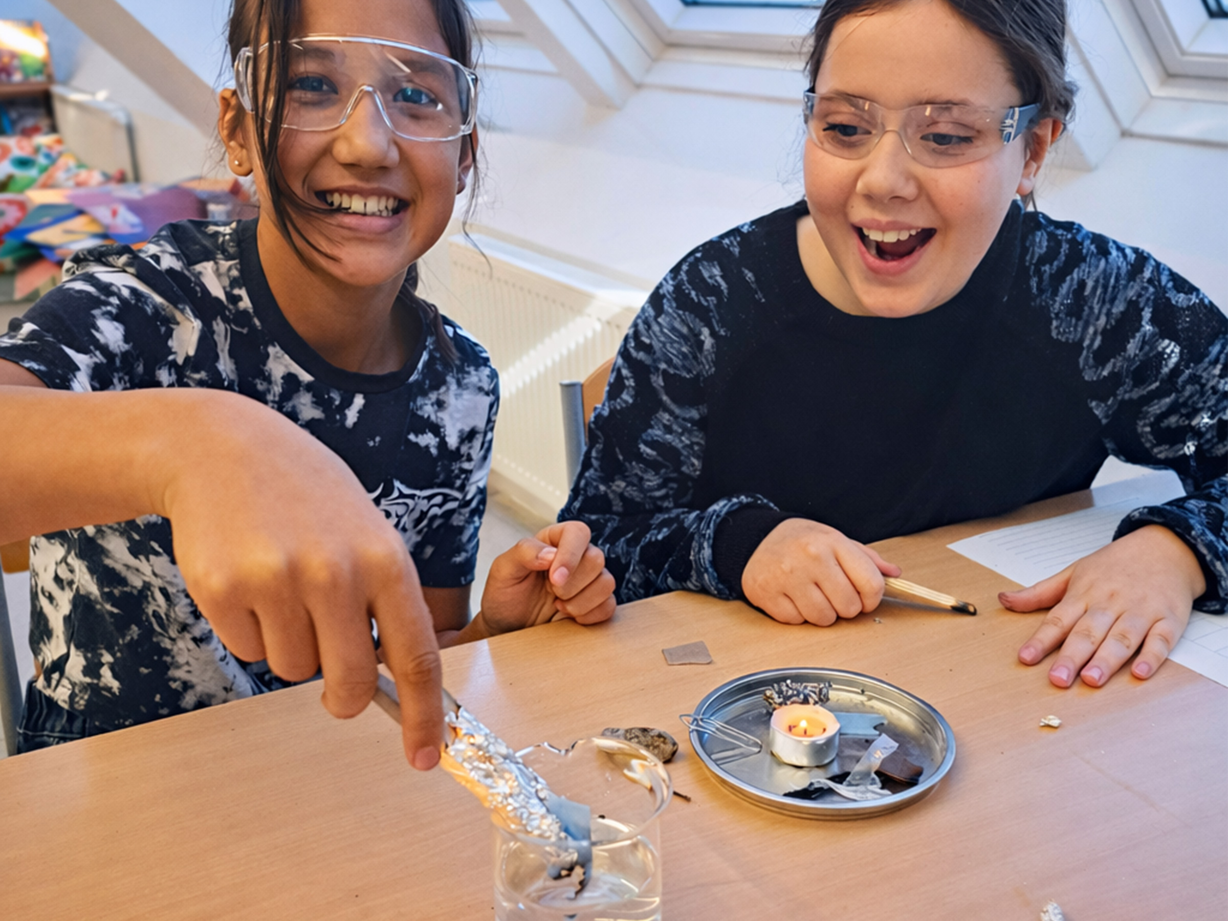 Zwei Mädchen mit Schutzbrille an einem Tisch sitzend mit lachendem und überraschtem Gesicht an einem Experiment mit Kerze und einem Wasserglas arbeitend