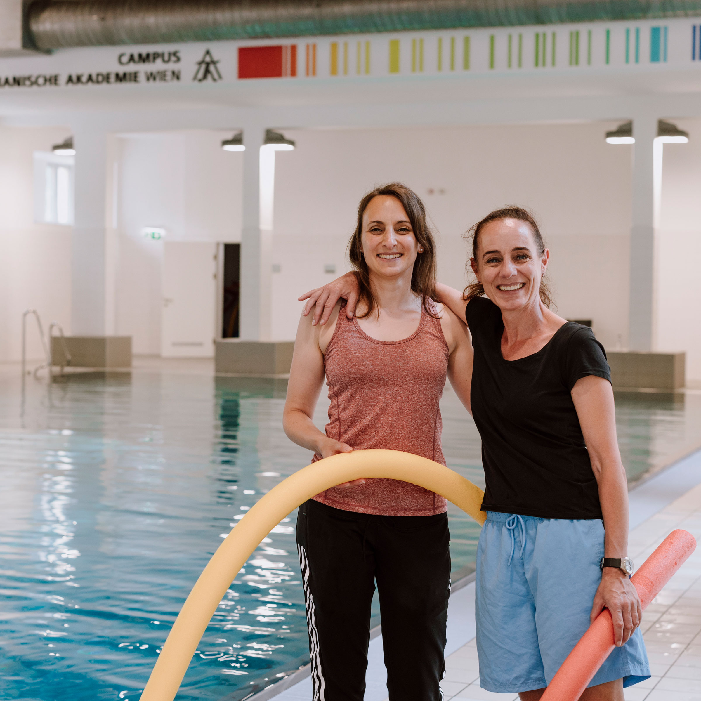 Zwei sportlich gekleidete Frauen, beide mit dunklen langen Haaren, und jeweils einer Schwimmnudel in der Hand in einem Hallenschwimmbad vor dem Becken stehend und in die Kamera lachend.