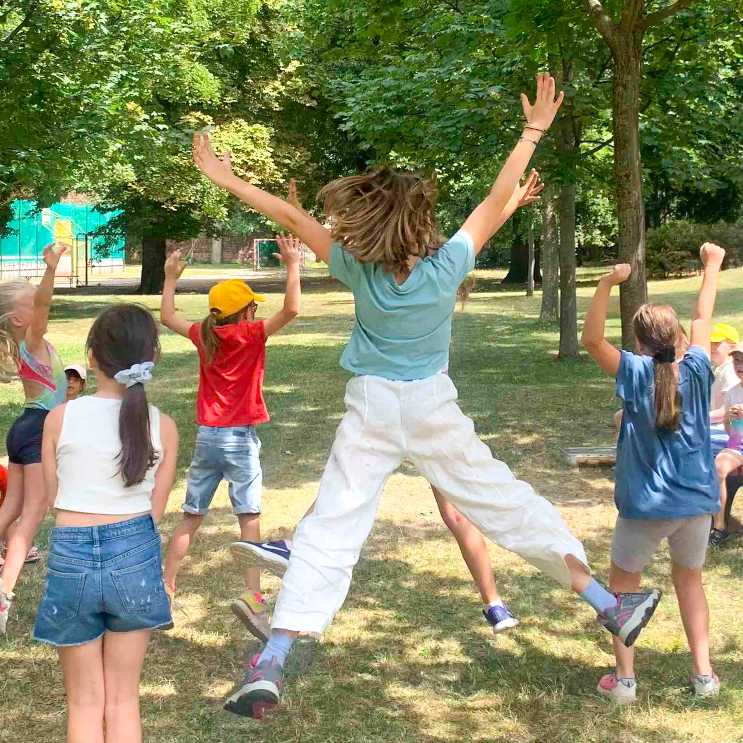 Mehrere Kinder in die Luft springend mit Armen in der Höhe in einm Park im Sommer in bunter Kleidung von hinten zu sehen