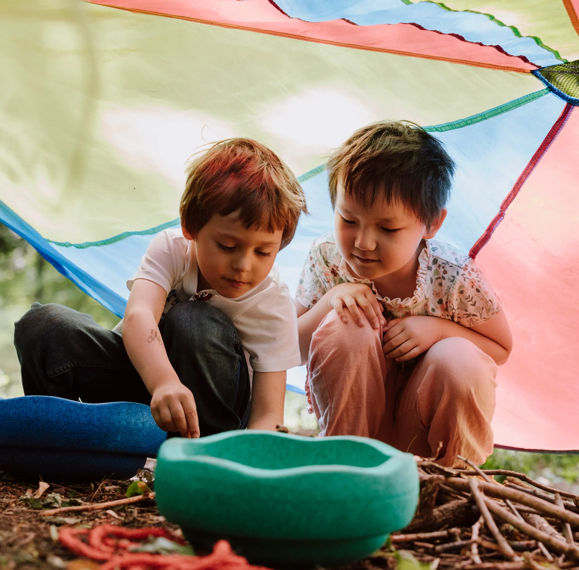 Zwei Kleinkinder ins Spiel vertieft am Waldboden unter einem bunten Sonnenschutz sitzend