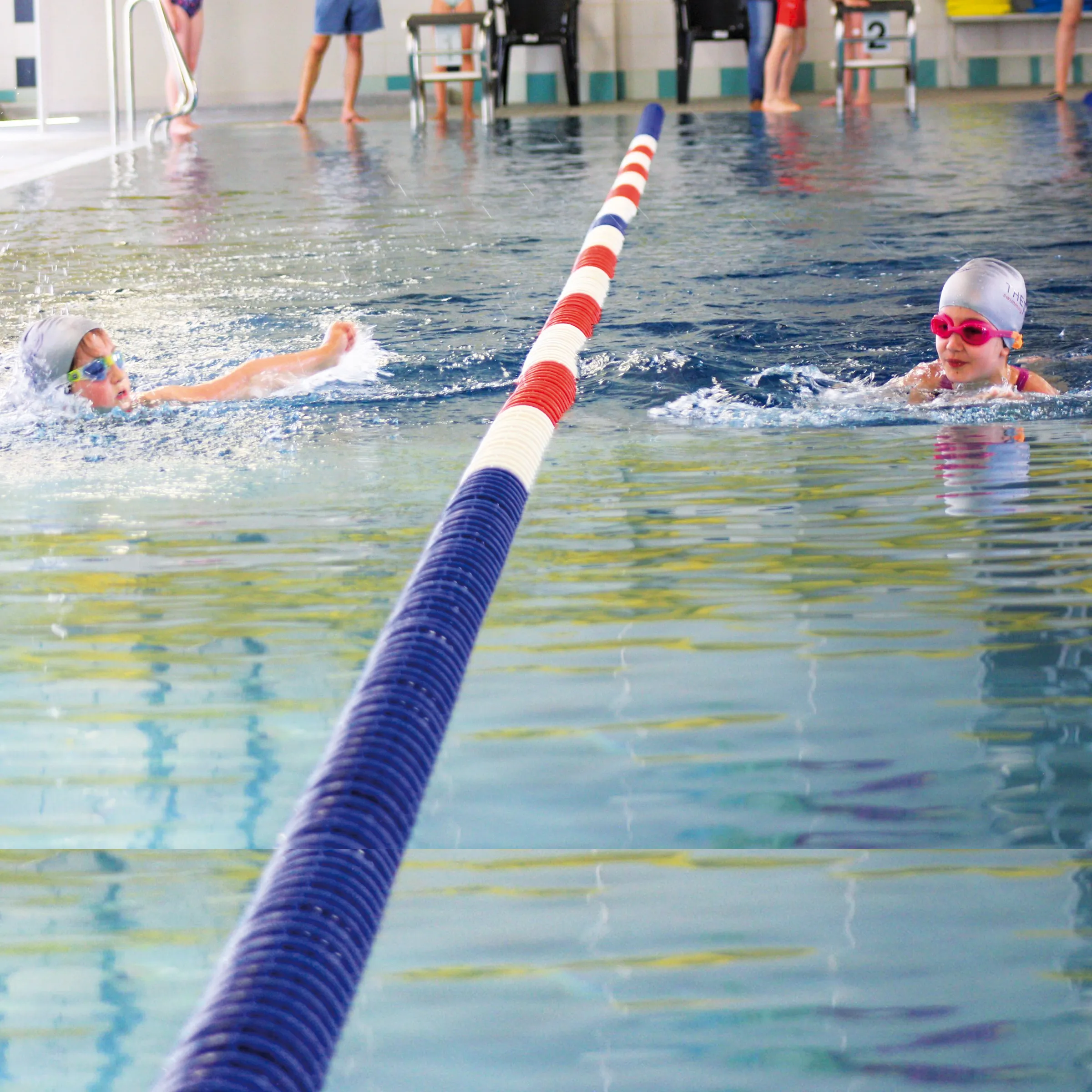 Zwei Kinder mit Badehaube und Schwimmbrille auf Bahnen in einem Hallenbad schwimmend