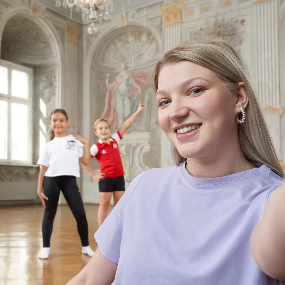 Junge Frau im lila T-Shirt vor einem barocken Hintergrund und einem Buben und einem Maedchen in Sportkleidung