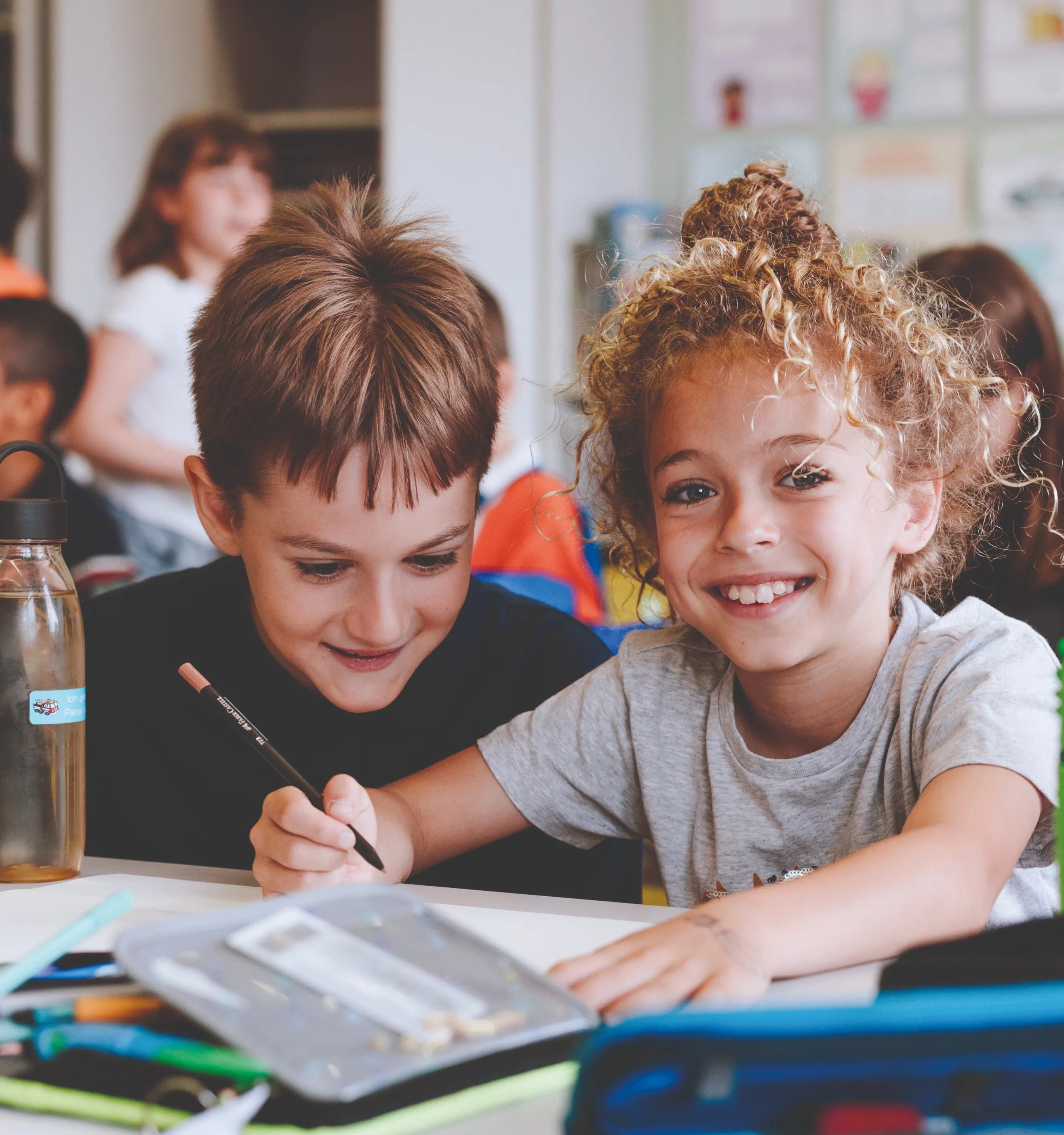 zwei Buben im Volksschulalter in der Schule an einem Tisch sitzend, einer nach unten blickend und schelmisch lachend, der andere in die Kamera blickend mit fröhlichem Lachen