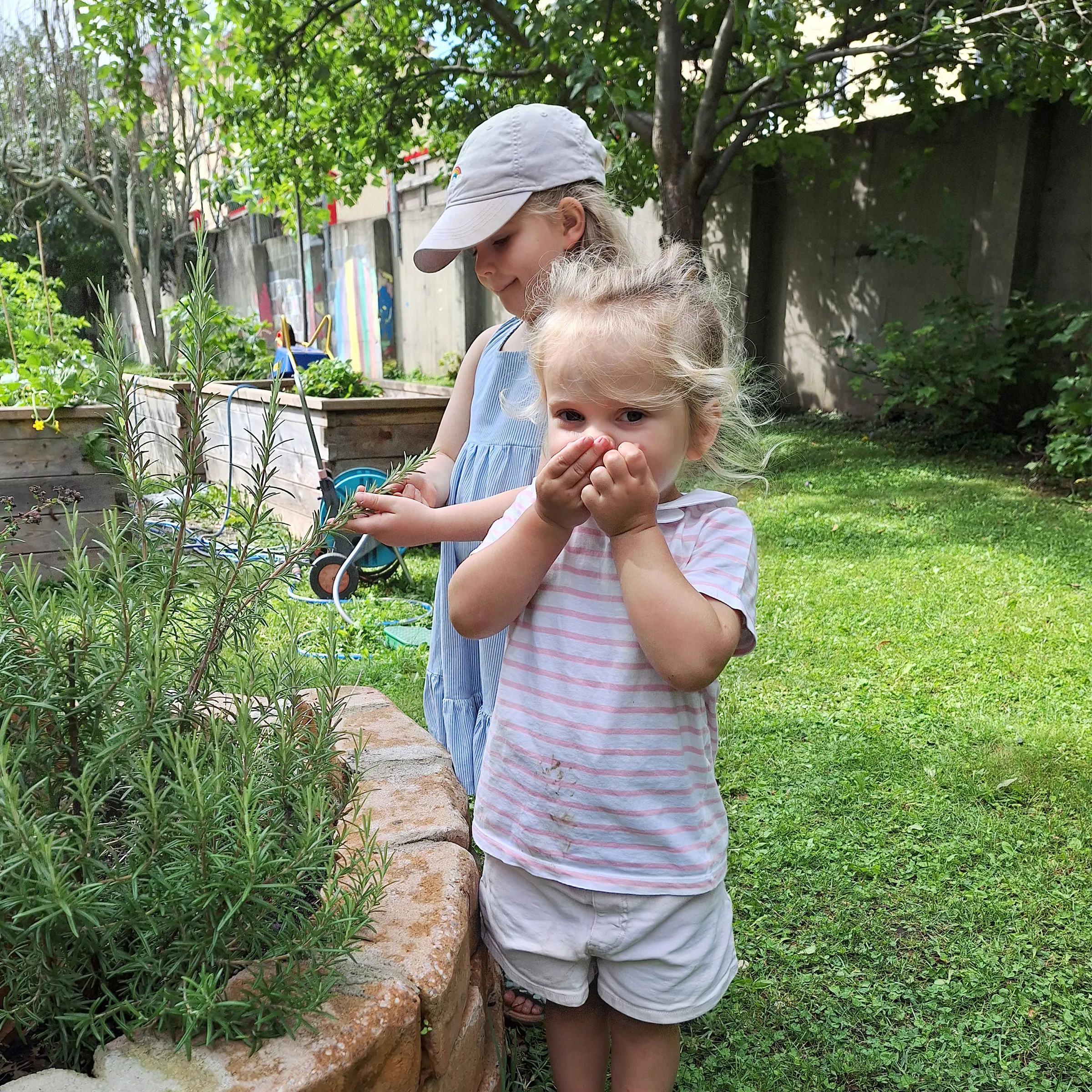Zwei kleine Mädchen in Sommerkleidung bei einer Kräuterspirale stehend, das vordere Kind an einem Stück Rosmarin riechend