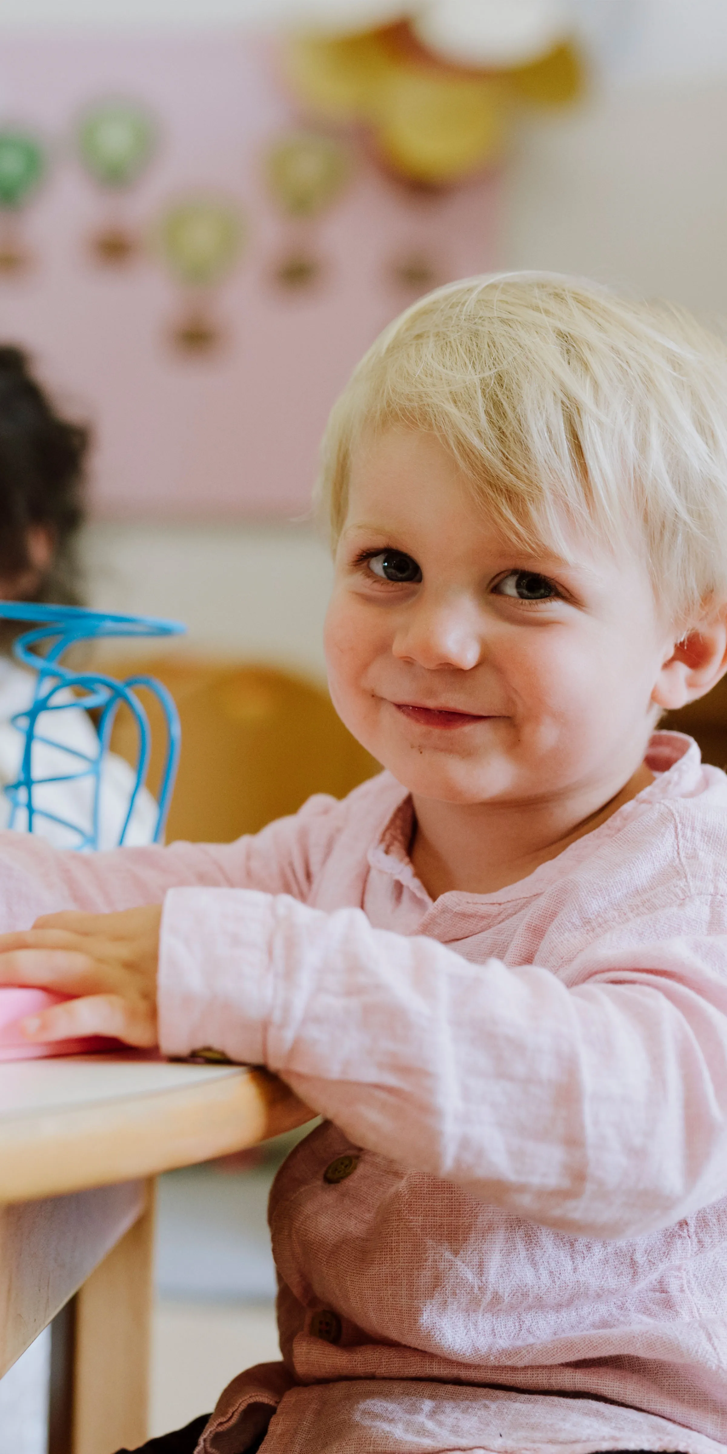 Kleiner blonder Bub an einem Tisch sitzend vor verschwommenem Hintergrund lächelt in die Kamera.