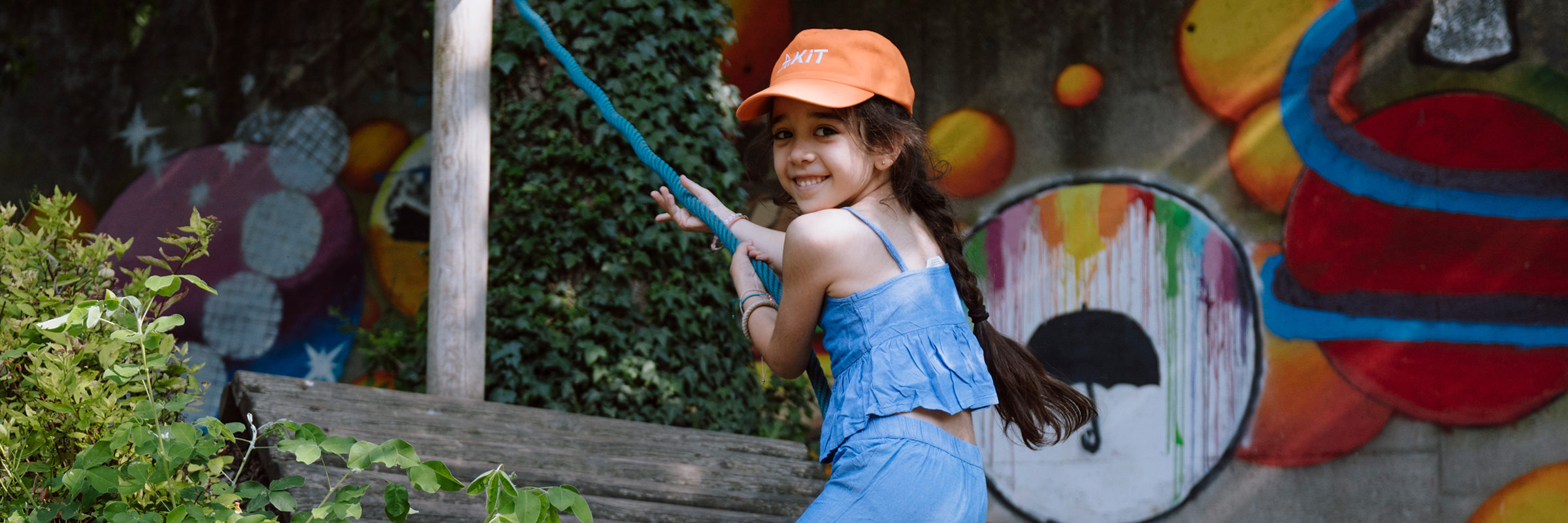 Kleines Mädchen mit dunklem Zopf und orange farbiger Kappe klettert im Freien an einem Seil eine schiefe Holzebene hoch.