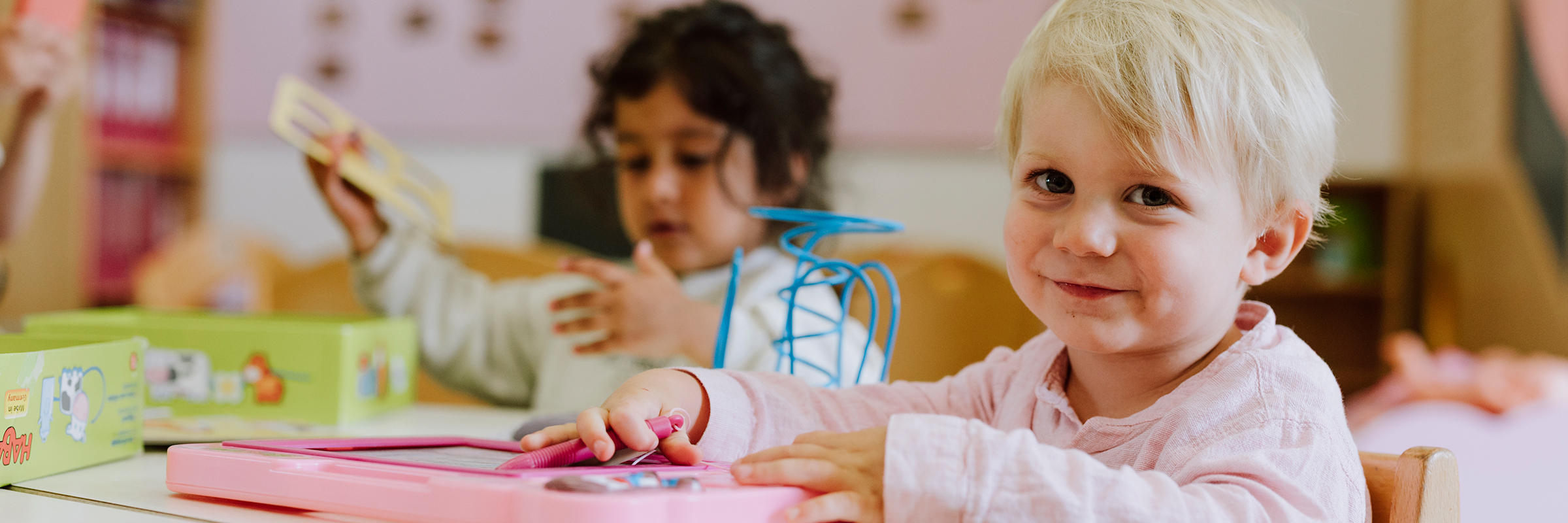 blonder kleiner Bub lacht freundlich in die Kamera und hält ein rosafarbenes Spielzeug in der Hand