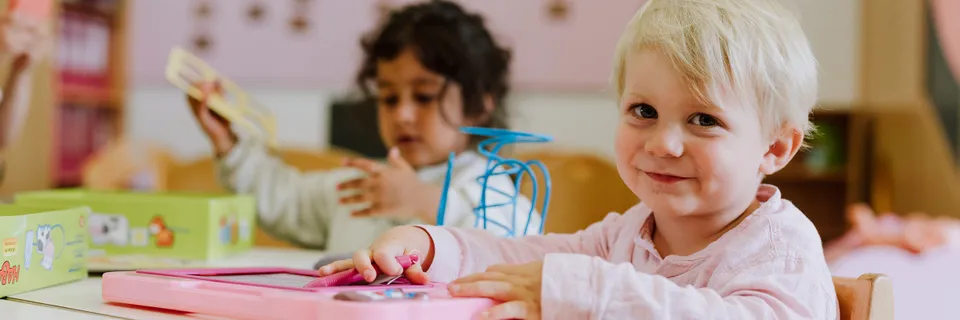 blonder kleiner Bub lacht freundlich in die Kamera und hält ein rosafarbenes Spielzeug in der Hand