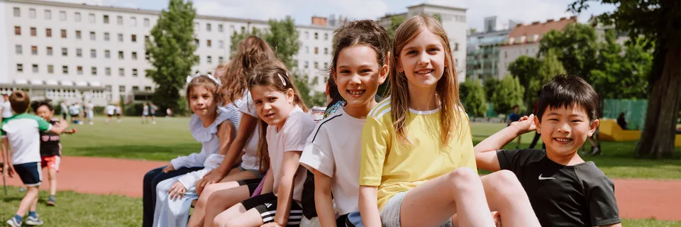 Eine Gruppe von Volksschulkindern in Sportkleidung am Sportplatz auf einer Bank sitzend und lachend in die Kamera grinsend