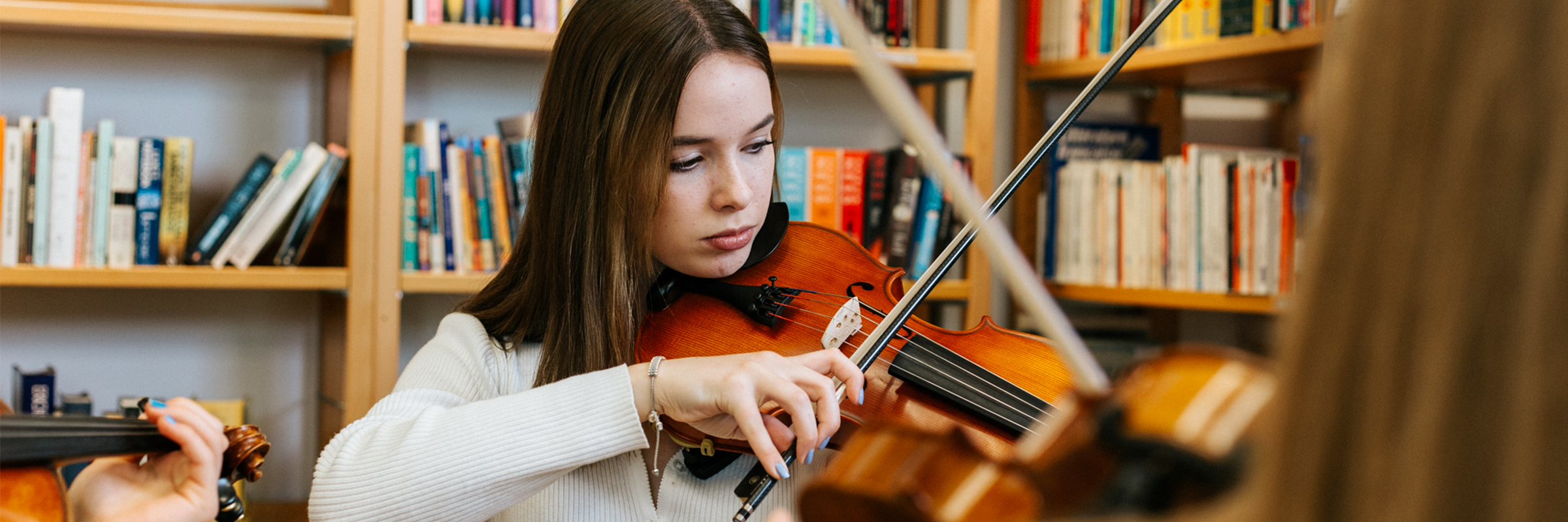 Junges Mädchen mit dunkelbraunen glatten langen Haaren vor einem Bücherregal Geige spielend