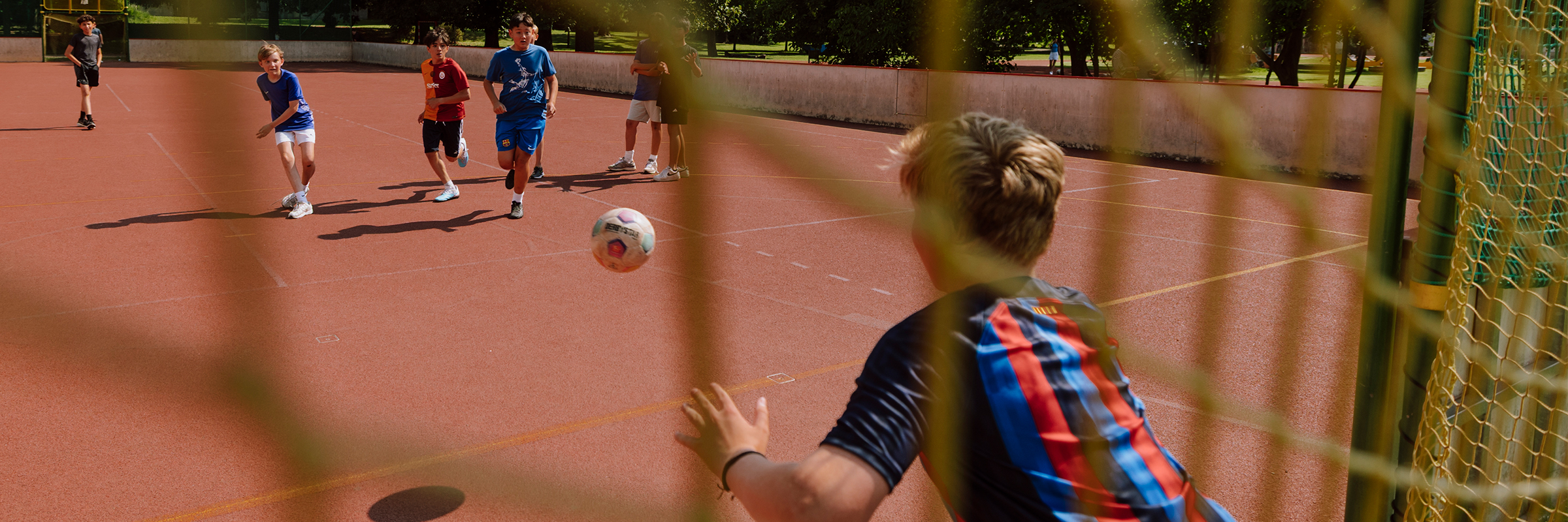 Hinter dem Tor auf einem Hartplatz, bei sonnigem Wetter dem Tormann, einem jungen Burschen, beim sieben Meter über die Schulter schauend