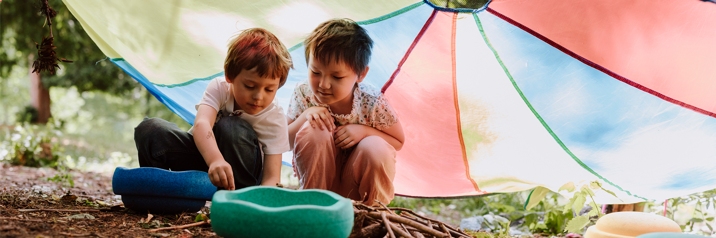 Zwei Kinder hocken auf einem Waldboden unter einem bunten Sonnedach und spielen konzentriert mit Holzstöckchen.