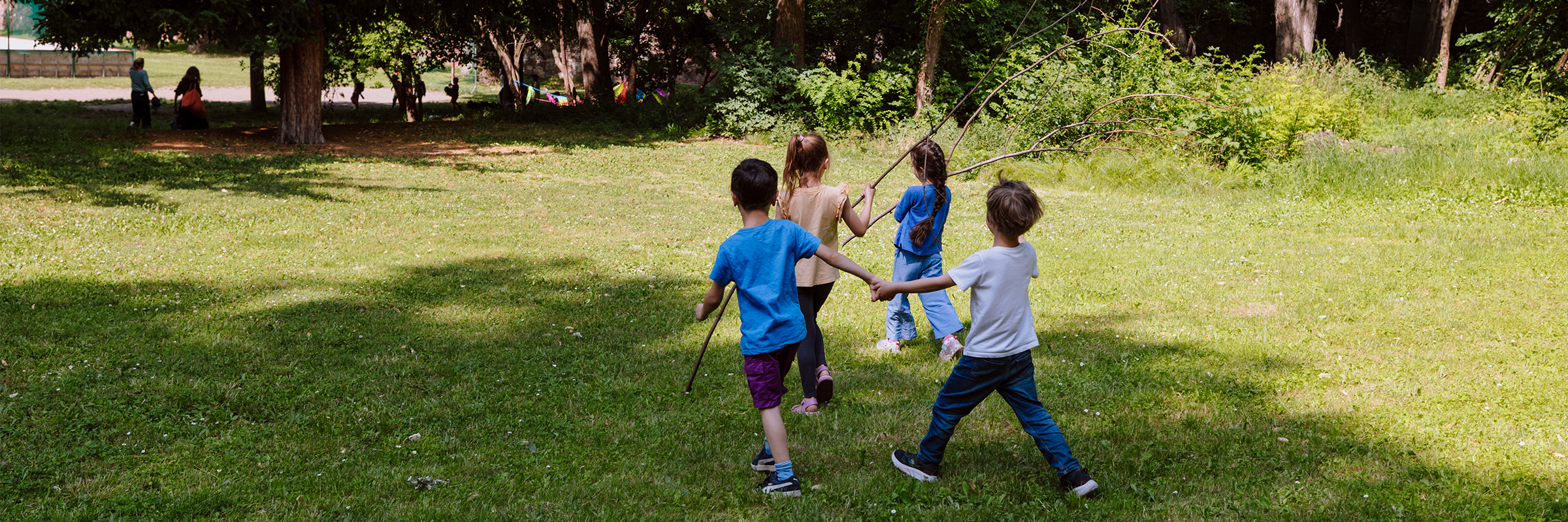 Vier Kinder von hinten aufgenommen laufen bei Sonnenschein im Sommer über eine Wiese, halten sich an den Händen. Drei von ihnen tragen auch lange Äste.