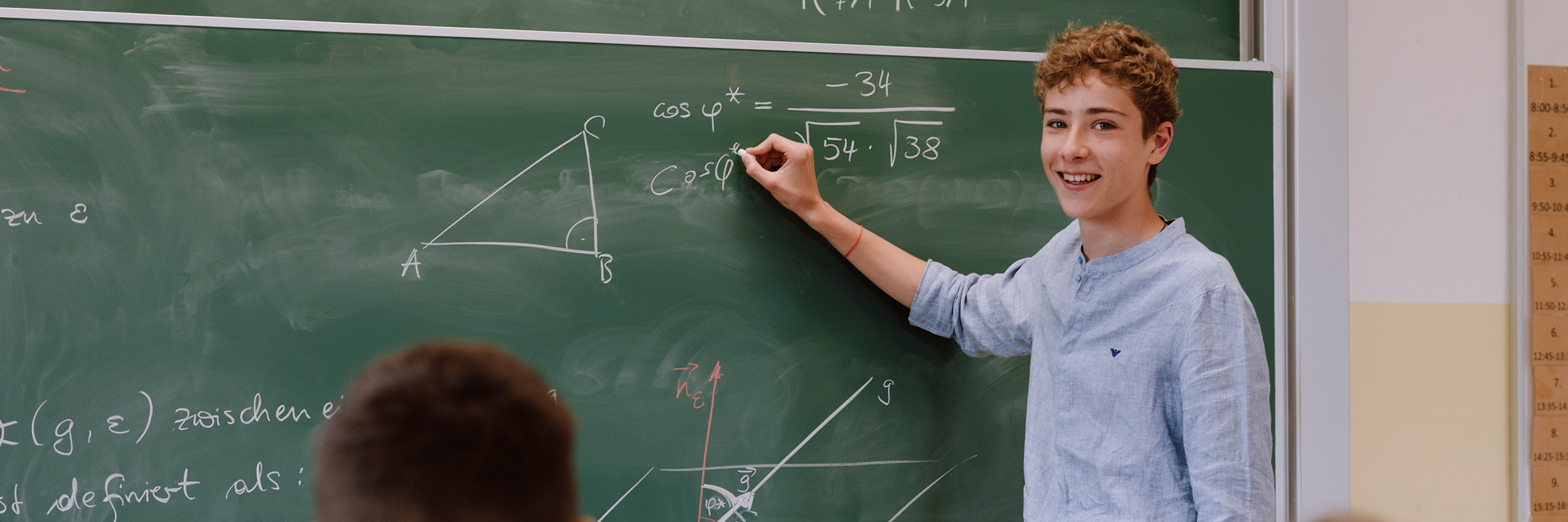 Junger Bursche mit blonden lockigen Haaren und in blauem Hemd an einer Schultafel mit Kreide stehend mit lachendem Blick in die Kamera eine mathematische Formel lösend
