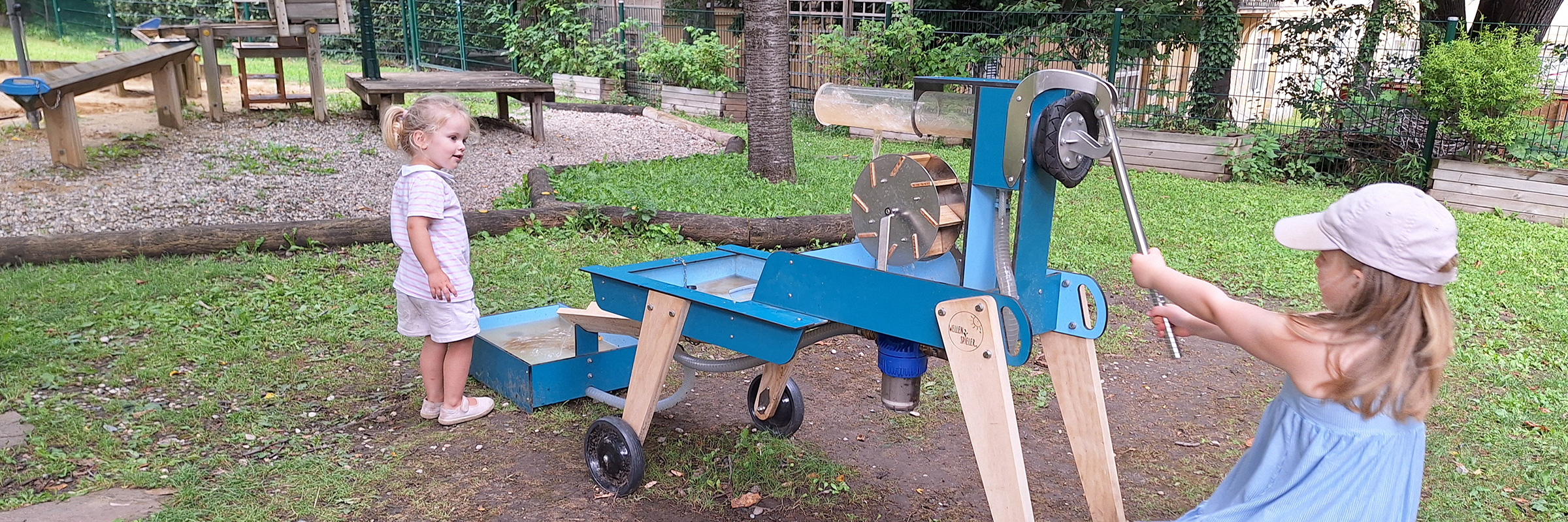 Zwei kleine Mädchen in luftiger sommerlicher Kleidung auf einem Spielplatz mit einer Wasserpumpe und einem Wasserrad beschäftigt