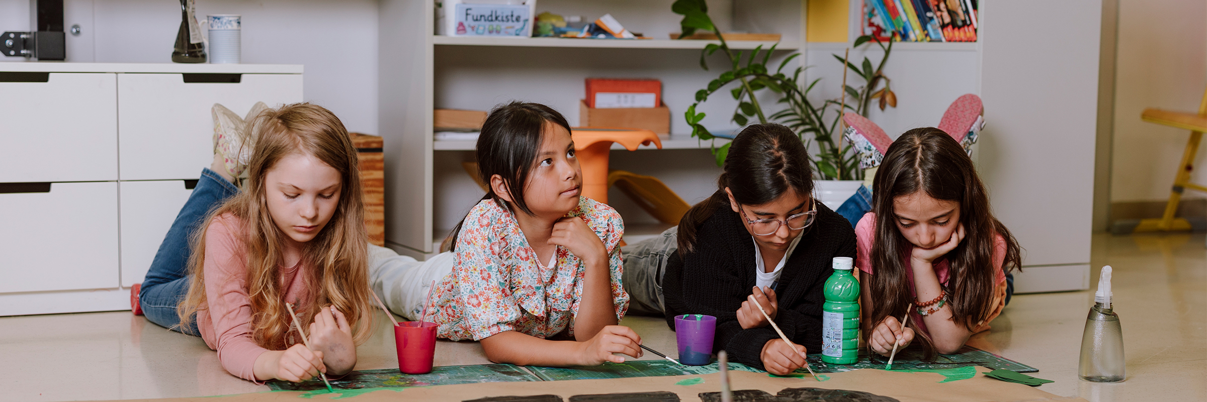 Vier Mädchen am Bauch liegend, malen ein übergroßes Plakat mit Pinsel und Wasserfarben