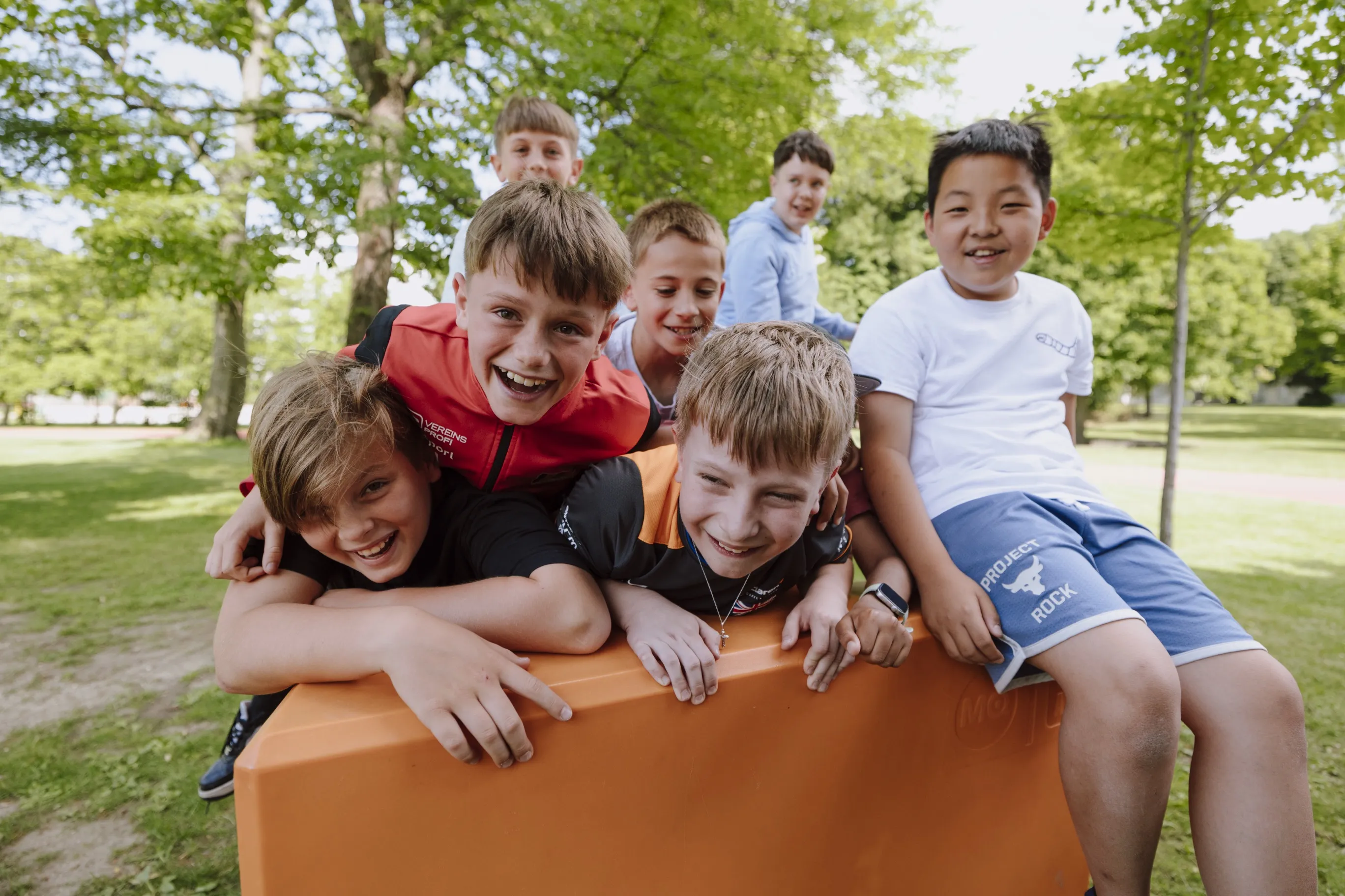 Gruppe von Buben in einem Park bei sommerlichem Wetter auf einem übergroßen orangefarbigen Sitzmöbel über- und nebeneinander liegend bei ausgelassener Stimmung