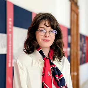 Portraitfoto einer jungen Frau mit Brille und schulterlangen braunen Haaren, weißer Bluse und rot blau weißem Seidenhalstuch vor einem historischen Hintergrund.