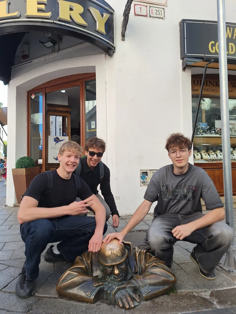 Three young men crouch next to a bronze statue of a man emerging from a manhole, posing and smiling at a street corner.
