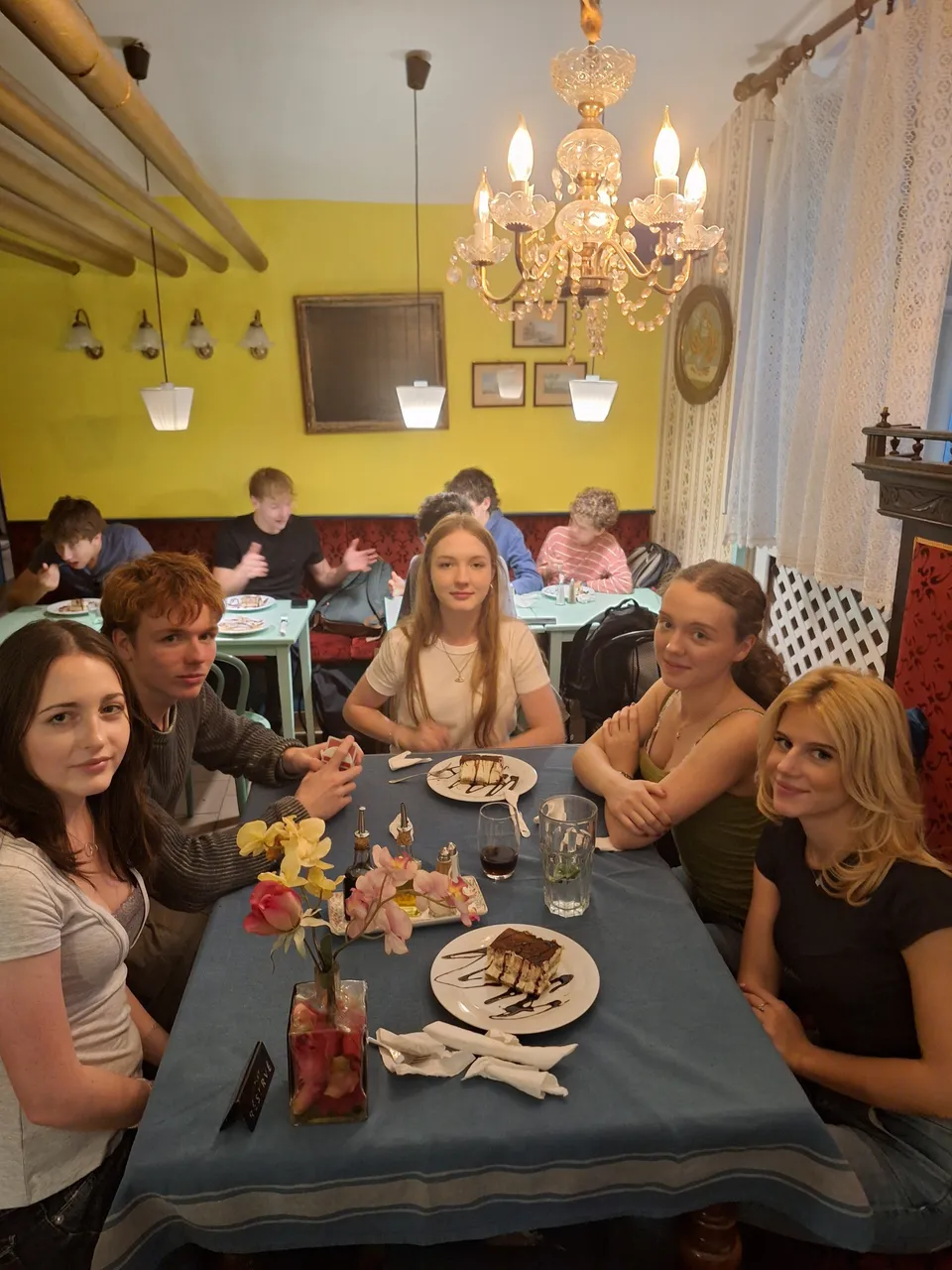 Five people are seated around a table in a cozy restaurant, sharing desserts. A chandelier hangs overhead, and other diners are seen in the background.