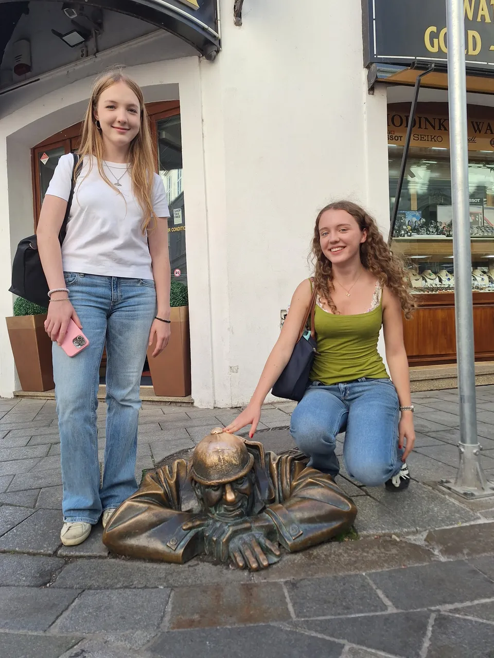 Two people pose next to a bronze statue of a man emerging from a manhole on a city street.