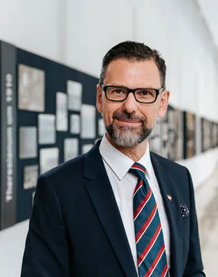 eleganter Mann mit dunklen Haaren, Vollbart, dunkler Brille und Anzug mit rot blauer Krawatte